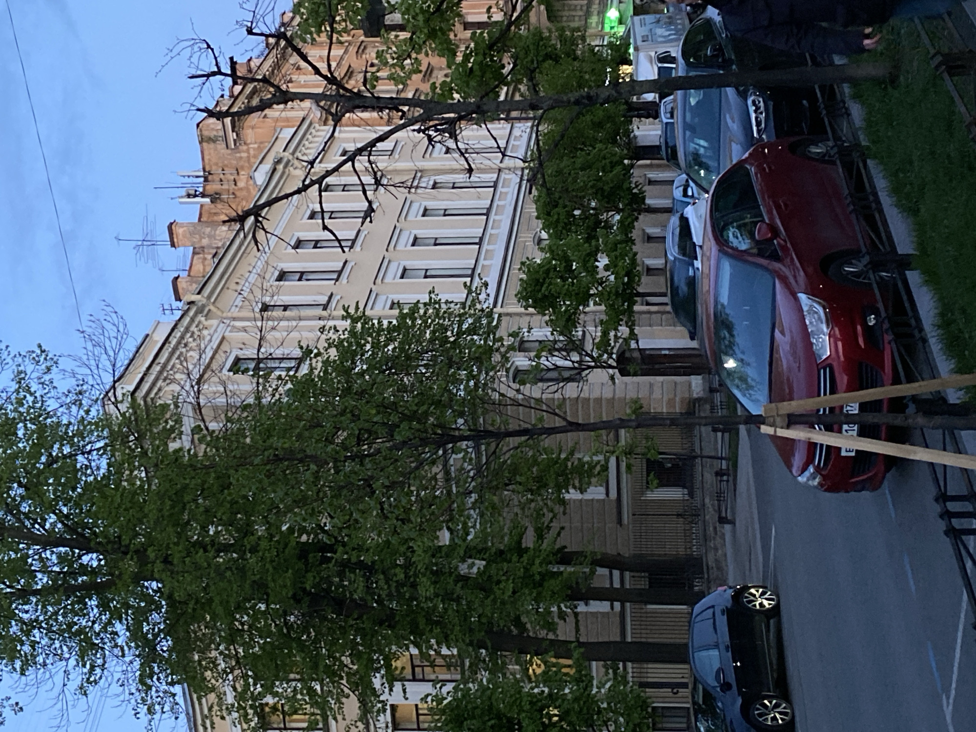 The image depicts a street scene in Saint Petersburg, Russia. A row of parked cars, including a red Ford, lines the street. Behind the cars is a sidewalk with a metal fence separating it from the road. Lush green trees partially obscure a multi-story building with ornate facade and windows. The sky is visible in the upper left, appearing to be early evening. There are people visible near the top right edge of the frame.