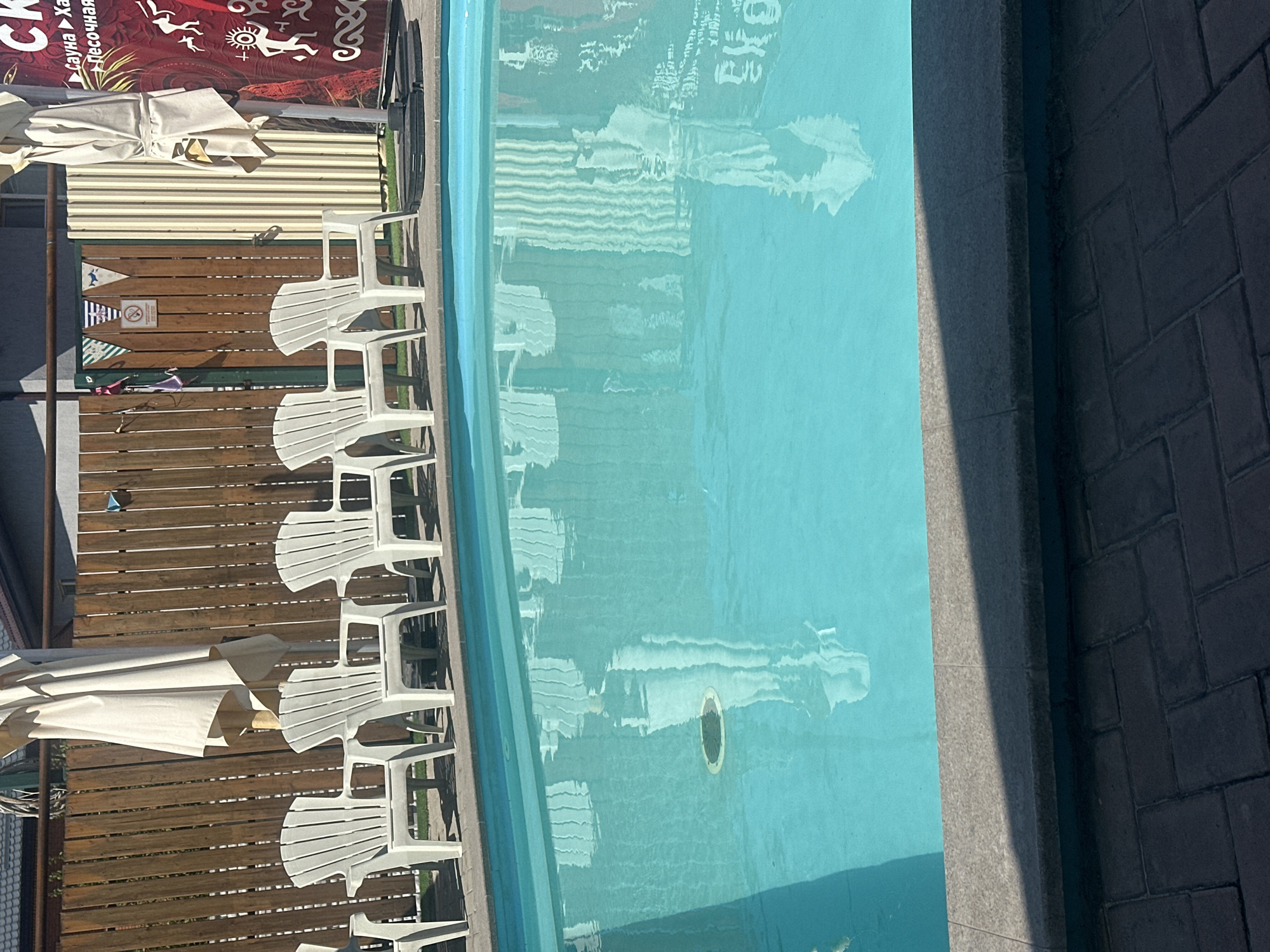 A bright blue swimming pool is located outdoors in Besagash, Kazakhstan. Several white plastic lounge chairs are arranged along the edge of the pool, facing the water. A tall, beige umbrella is positioned behind the chairs. In the background, a wooden fence runs horizontally, with a vertical structure of light-colored corrugated metal to its right. A red banner with white text and graphics is also visible on the far right. The pool water reflects the surrounding environment, including the chairs and the corrugated metal structure. The ground around the pool is paved with dark grey bricks.