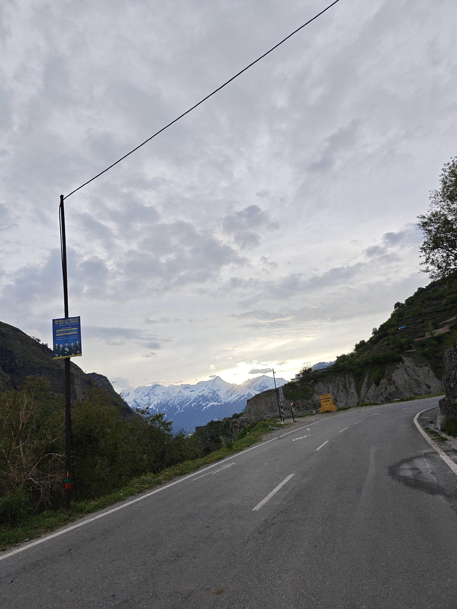 A paved road curves to the right in Khangsar, India, under a cloudy sky. Snow-capped mountains are visible in the distance. A pole on the left side of the road displays a sign for Kullu Valley School, advertising admissions. A power line extends diagonally across the upper part of the frame. Vegetation covers the hillsides on either side of the road. A yellow sign is visible on the right side of the road. A small patch of water is on the right edge of the road.