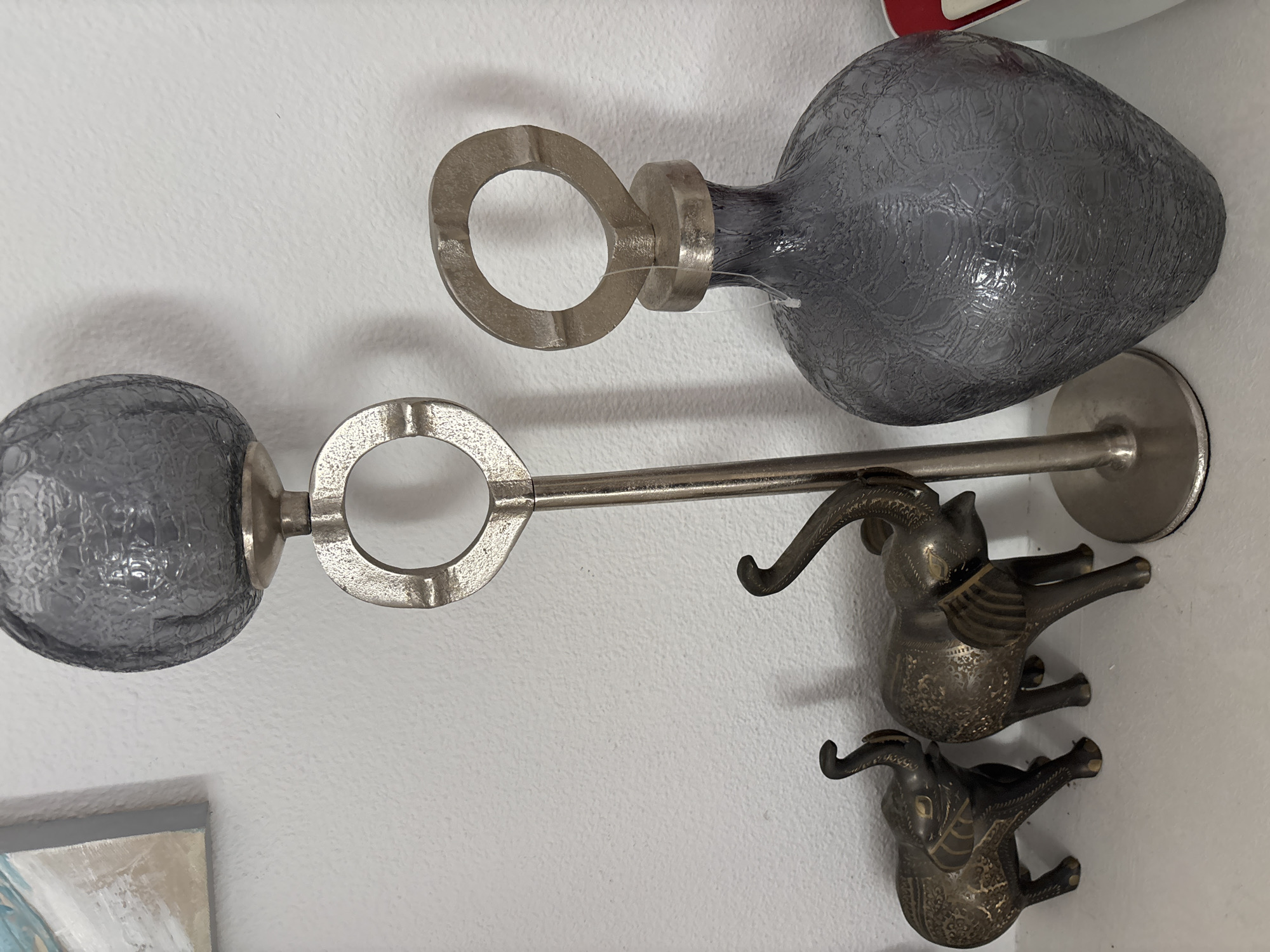 This image depicts a still life arrangement of decorative objects on a white shelf against a white textured wall. The arrangement includes two small, dark metal elephant figurines with detailed etchings, positioned on the left. To the right of the elephants is a tall, slender silver candelabra with a circular opening in its stand and a textured dark gray globe at the top. Further to the right is a dark gray textured glass bottle with a silver cap that has a circular handle. A portion of a painting with abstract blue and beige elements is visible on the upper left edge of the image. The scene is indoors, likely in a home or shop. There are no people or animals present, and no actions are taking place. The environment is clean and organized.