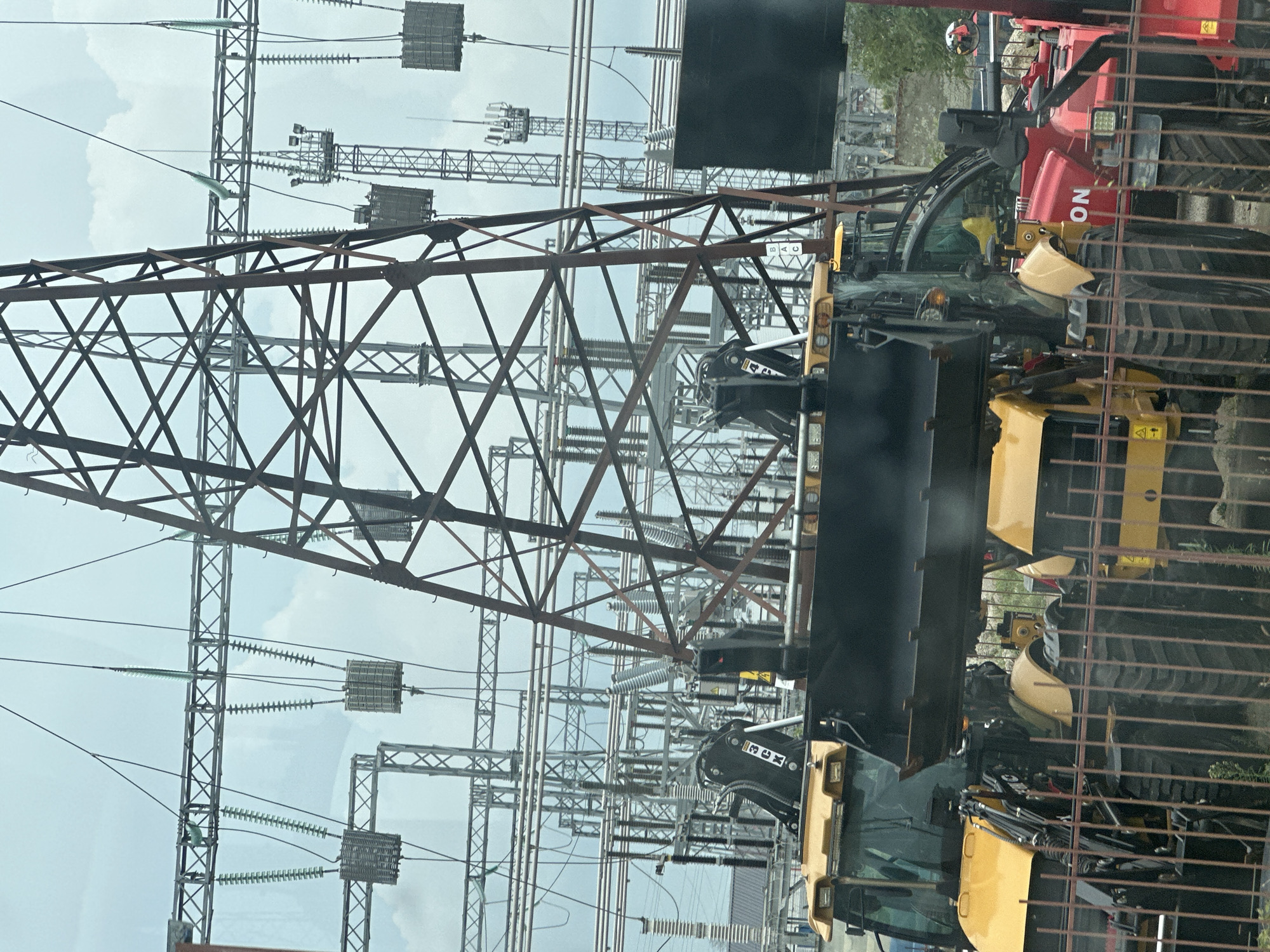 The image depicts an outdoor scene at an electrical substation in Beryozovy, Russia. In the foreground, behind a metal fence, are several yellow construction vehicles, likely backhoe loaders, with their buckets raised. One vehicle has the text "3CK" visible on its arm. To the right of the yellow vehicles, a red construction vehicle is partially visible, with the word "ON" prominently displayed on its side. In the background, a complex network of electrical towers, power lines, and insulators dominates the landscape under a cloudy sky. The overall impression is one of industrial infrastructure and heavy machinery.