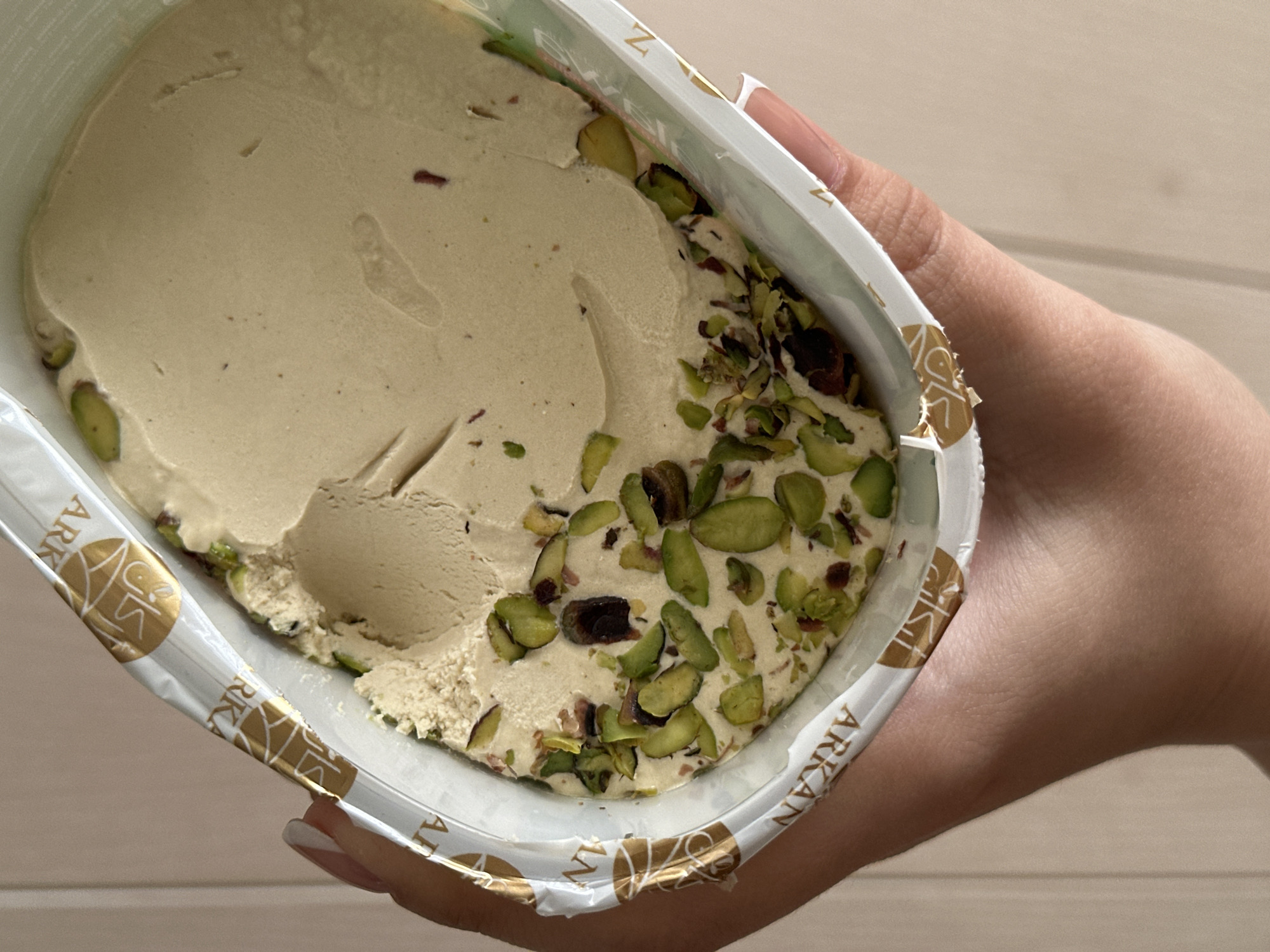 A human hand, featuring a manicured nail with a French tip, holds a rectangular plastic container of halva. The container's rim displays a white and gold foil label with the brand name "ARKAN" visible. Inside, a light cream-colored halva is present, with its surface appearing disturbed and textured on the left side, while remaining relatively smooth on the right. Numerous green pistachio pieces and scattered reddish-brown fragments are embedded within and atop the halva, particularly concentrated in the lower and left sections. The background is an indistinct, light-toned surface featuring a vertical line, possibly a wall or doorframe. The scene is located in the city of Chaikovsky, Russia.