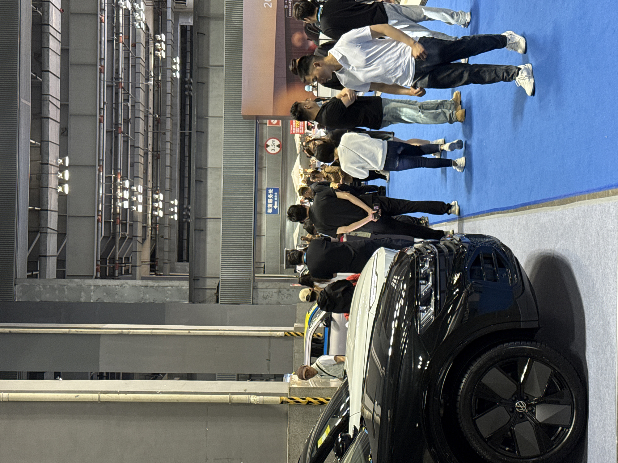 The image captures a scene at a car exhibition located in an interior setting, likely a convention center or large indoor arena in an unknown city. A black Volkswagen car is prominently displayed on a grey carpeted floor, and a crowd of people are gathered nearby. The people appear to be casually dressed, and some are wearing masks. Overhead, there is an industrial ceiling with numerous pipes and lighting fixtures. Behind the crowd, a blue carpet area is visible, and additional exhibition displays can be seen in the background. A sign is visible on a pillar.