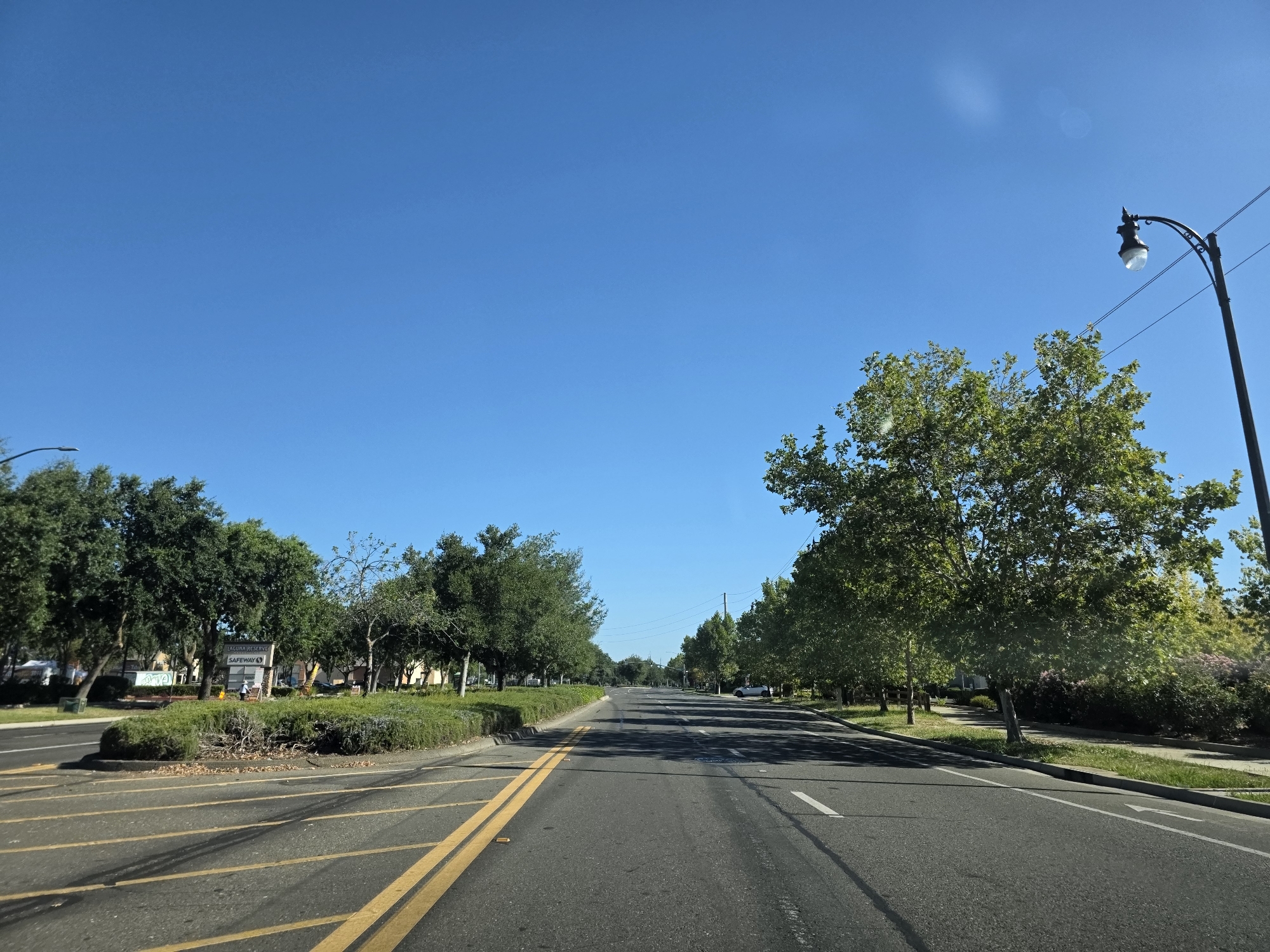 The image depicts a clear blue sky over a suburban street in Elk Grove, California. The perspective is from a moving vehicle, looking down a two-lane road lined with trees. On the left side of the road, there is a landscaped median with a "Laguna Reserve" sign and a "Safeway" store visible in the background. The road has yellow double lines indicating traffic separation and white lane markers. A street lamp is visible on the right side of the image, with power lines stretching across the sky. The trees are lush and green, suggesting a temperate climate. No people are clearly visible in the image. The overall environment is peaceful and well-maintained, typical of a residential or commercial area.