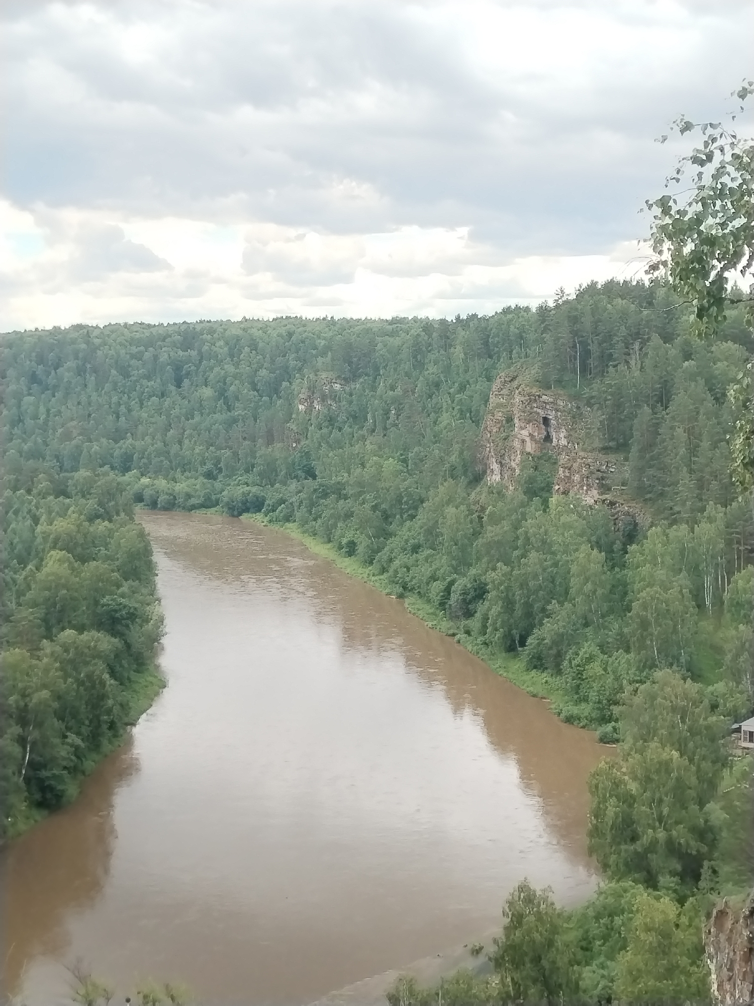 The image captures a scenic view of the Usva River as it flows through a forested landscape near Idrisovo, Russia. The river is wide and murky, with a light brown hue, indicating potentially high water or sediment content. It winds its way through a dense forest that covers the rolling hills on either side. The vegetation is predominantly deciduous trees with lush green foliage, creating a thick canopy. On the right bank, a prominent rock formation rises from the forest, its weathered surface showing signs of erosion and a dark opening that could be a cave or crevice. The sky is overcast with a layer of grey clouds, suggesting a mild or possibly rainy day. A small, rustic wooden building with a dark roof is partially visible nestled amongst the trees on the right bank, near the river's edge. The overall impression is one of a remote and natural environment.