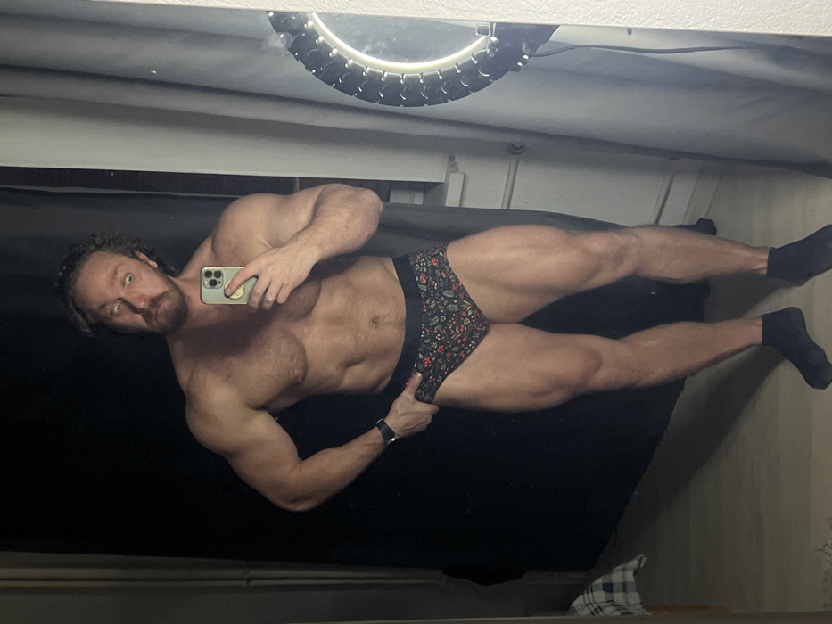 A muscular man is captured in a mirror selfie, standing shirtless and wearing brief-style underwear with black ankle socks. He has light skin, dark curly hair, a beard, and a mustache. His eyes are wide open, looking directly into the camera lens of the smartphone he holds in his right hand. The phone is light-colored and has a grip accessory on its back. His left hand is resting near his left hip, lightly touching the patterned underwear. A dark watch is visible on his left wrist.
The man exhibits a strong, athletic build with defined chest muscles, visible abs, and well-developed arms and legs. His underwear is dark with a vibrant, intricate pattern of small red berries and green leaves.
The setting appears to be an indoor space, likely a private room. The mirror reflects a dark background directly behind the man. Above his head in the reflection, a circular light fixture with individual glowing elements is visible, possibly a ring light or vanity light. The ceiling or upper wall area reflected in the mirror is a light color. At the bottom of the reflected image, a light-colored floor and a piece of plaid fabric or towel are discernible. The overall lighting suggests an artificial light source, likely from the visible ring light. The image itself is rotated 90 degrees counter-clockwise.