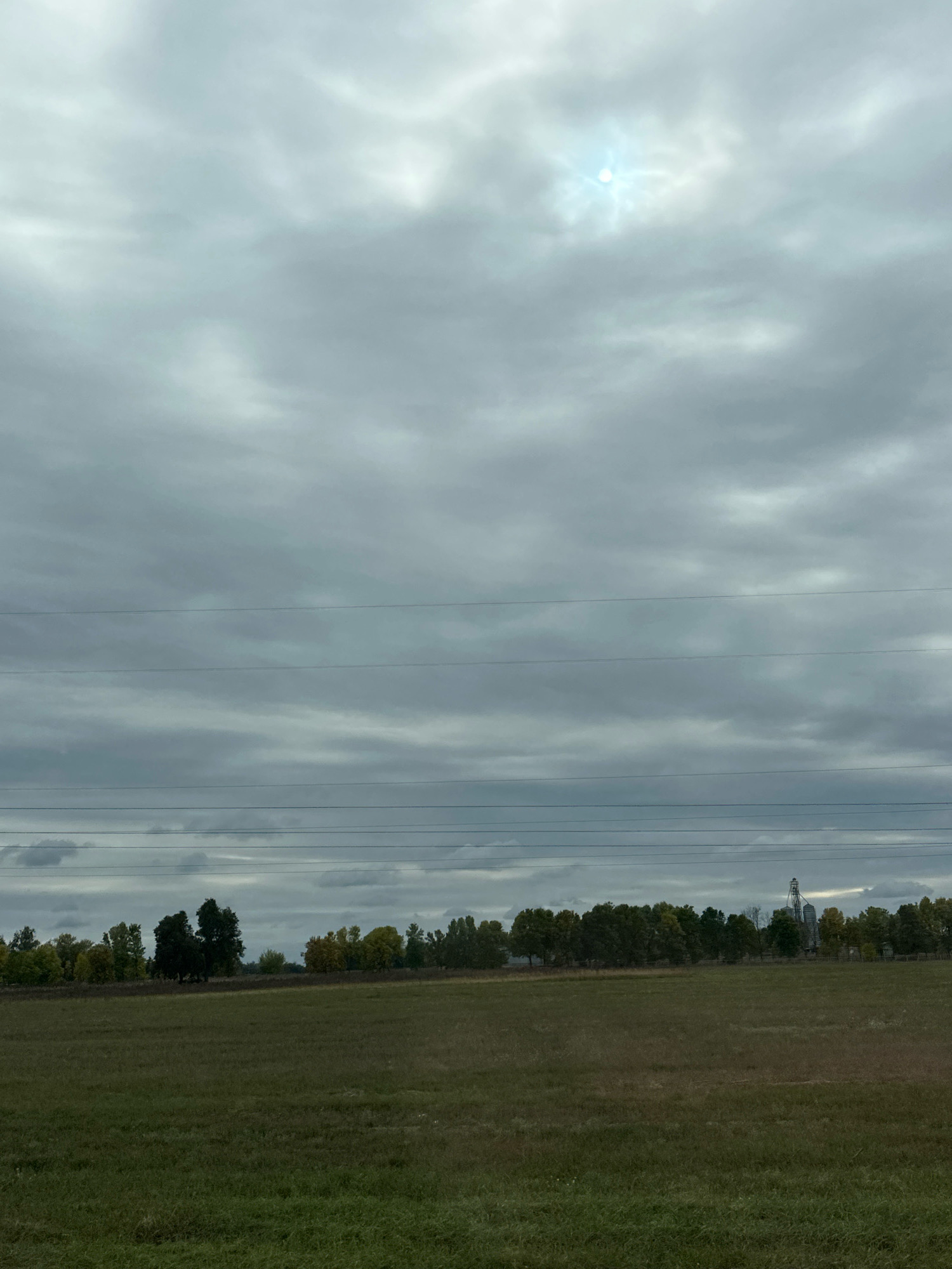 The image depicts an overcast sky with a faint sun peeking through the clouds. Below the sky, a line of trees with autumnal foliage stretches across the horizon. In the foreground, a vast, green field is visible. Several power lines are strung across the scene, dividing the sky and the landscape. To the far right of the tree line, a silo structure can be seen, suggesting a rural or agricultural setting. The overall impression is a tranquil, natural landscape under a moody sky, likely located in the countryside of Парыцкі сельскі Савет, Belarus.
