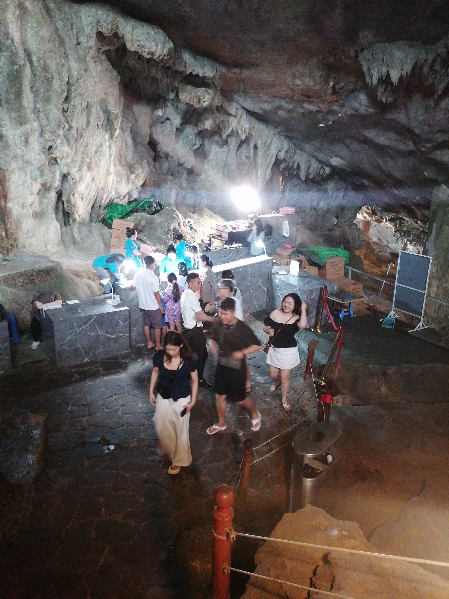 The image depicts a bustling scene inside a large natural cave, likely a tourist attraction in Bai Chay Ward, Vietnam. The cave features impressive limestone formations, including textured walls and a ceiling with numerous stalactites. The ground is paved with dark, irregular stones, forming a clear pathway for visitors.
Numerous people are present, indicating a busy period. In the mid-ground, a group of individuals, some wearing light blue shirts, appear to be staff members, possibly assisting visitors at two dark, marbled counters. Tourists, including adults and a few children, are seen walking, standing, and interacting within the cave. A woman in a black off-shoulder top and white skirt is prominently featured in the foreground, smiling as she moves towards the left. Another woman in a long white skirt walks further left, away from the viewer.
Artificial lighting, particularly a bright source in the mid-background, illuminates the cave's interior. Scattered around the counters are green tarps, cardboard boxes (one with partially visible text like "MIA" or "MIAO"), and small stools. To the right, a white-framed blackboard stands near the cave wall, alongside a blue dustpan and brush. Red stanchions with ropes guide the flow of people down a slight incline on the right, where a silver trash can is also visible. The setting clearly functions as an organized tourist site.