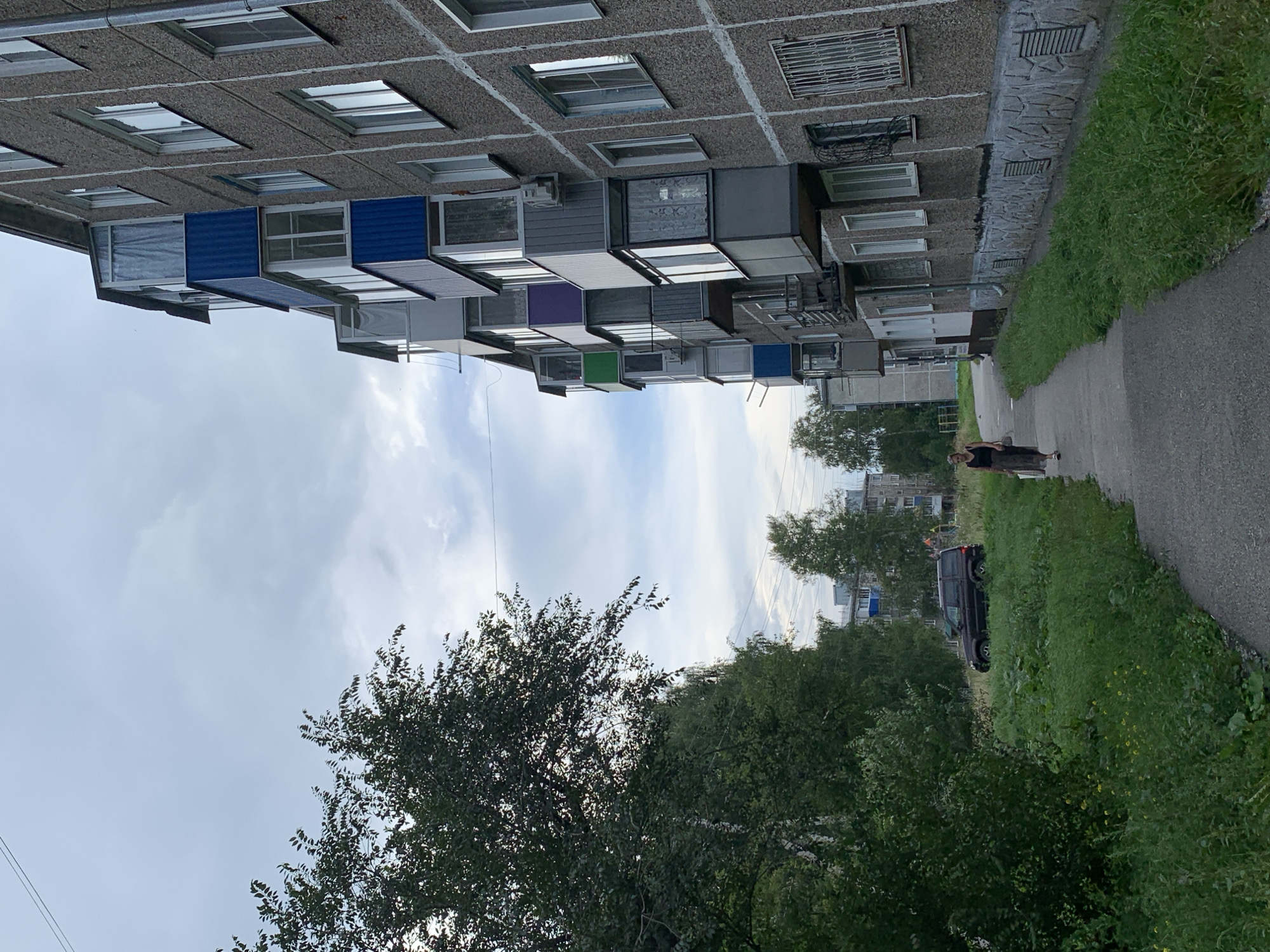 A woman walks along a paved path next to a large, multi-story apartment building in Prokopyevsk, Russia. The building's exterior is rough-textured concrete with a series of balconies, some of which are enclosed with colorful panels in blue, purple, and green. The ground is covered with green grass and some weeds. To the left of the path, there is a dense cluster of trees and a dark-colored SUV parked in a grassy area. The sky is overcast with clouds.