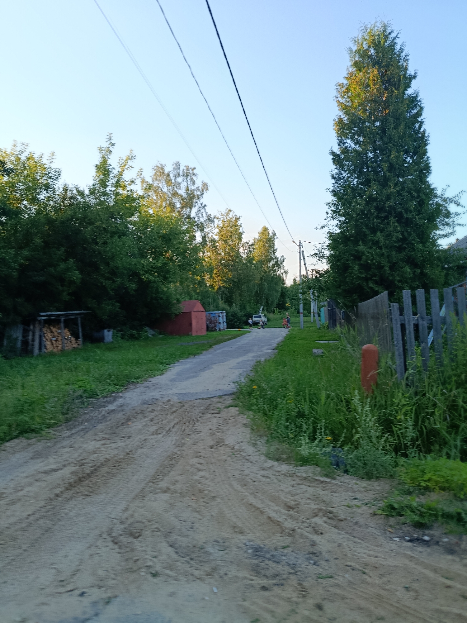 The image depicts a rural road in Vereya, Russia. The road is unpaved and covered in sand with visible tire tracks. On the left side of the road, there is a stack of firewood under a simple wooden shelter, and a small red outbuilding. Further down the road, trees and greenery line the path. A tall, dark green evergreen tree stands prominently on the right side, accompanied by a weathered wooden fence. In the distance, a few people and a white vehicle are visible on the road. Power lines stretch across the clear blue sky. The overall scene suggests a quiet, possibly summer day in a village setting.