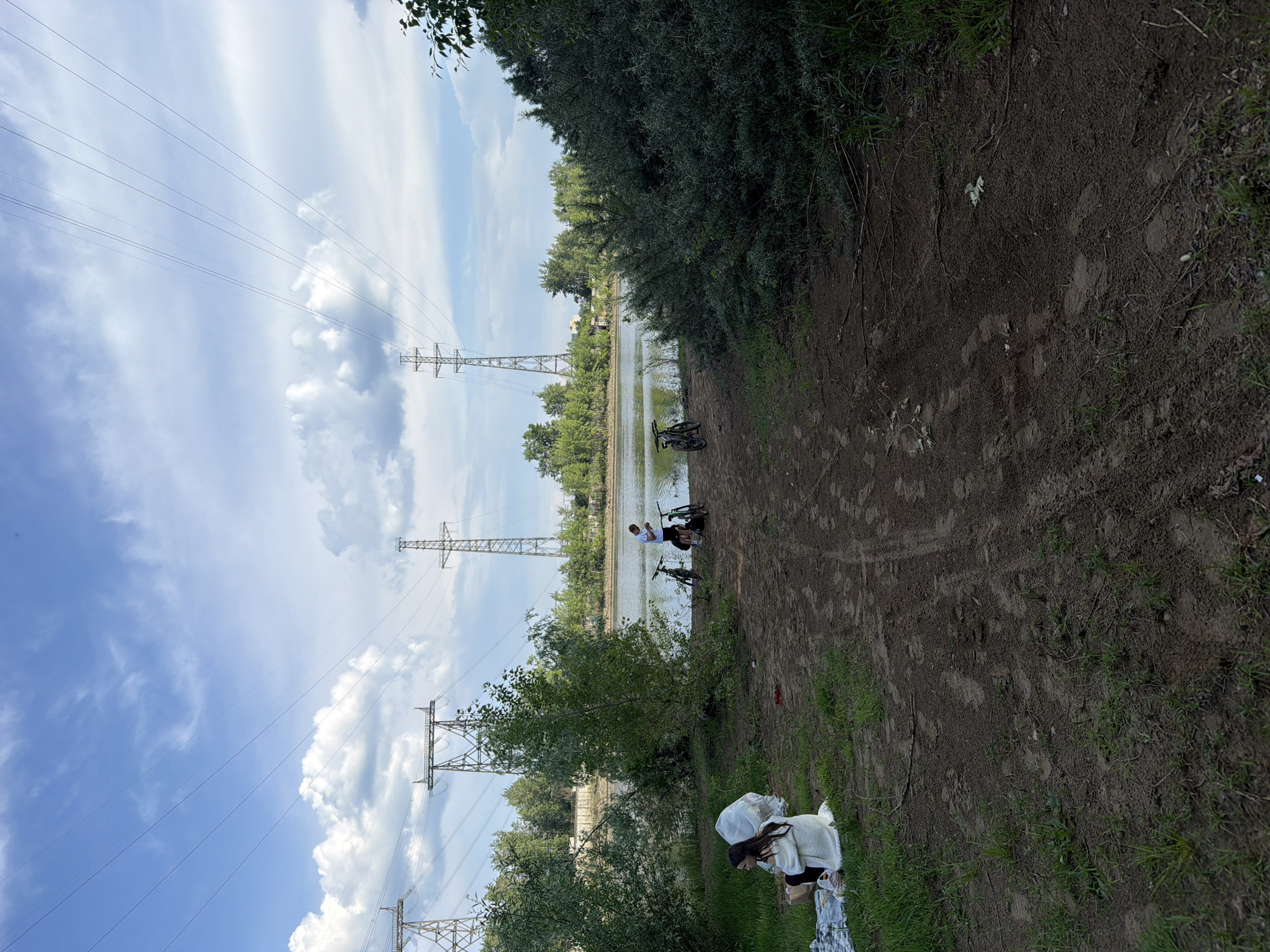 The image depicts a lakeside scene in Chaikovsky, Russia, likely during daylight hours given the bright sky. In the foreground, a young woman with dark hair is seated on a blanket, facing away from the camera, with white trash bags beside her. The ground is a mix of grass and dirt, with visible tire tracks and scattered debris. A body of water, presumably a lake or river, is visible in the mid-ground.
Across the water, a line of trees forms the background, with several tall, industrial power line towers scattered throughout the landscape. A man is standing near the water's edge, next to two bicycles. The sky is blue with scattered white clouds. The overall impression is of a recreational area near industrial infrastructure.