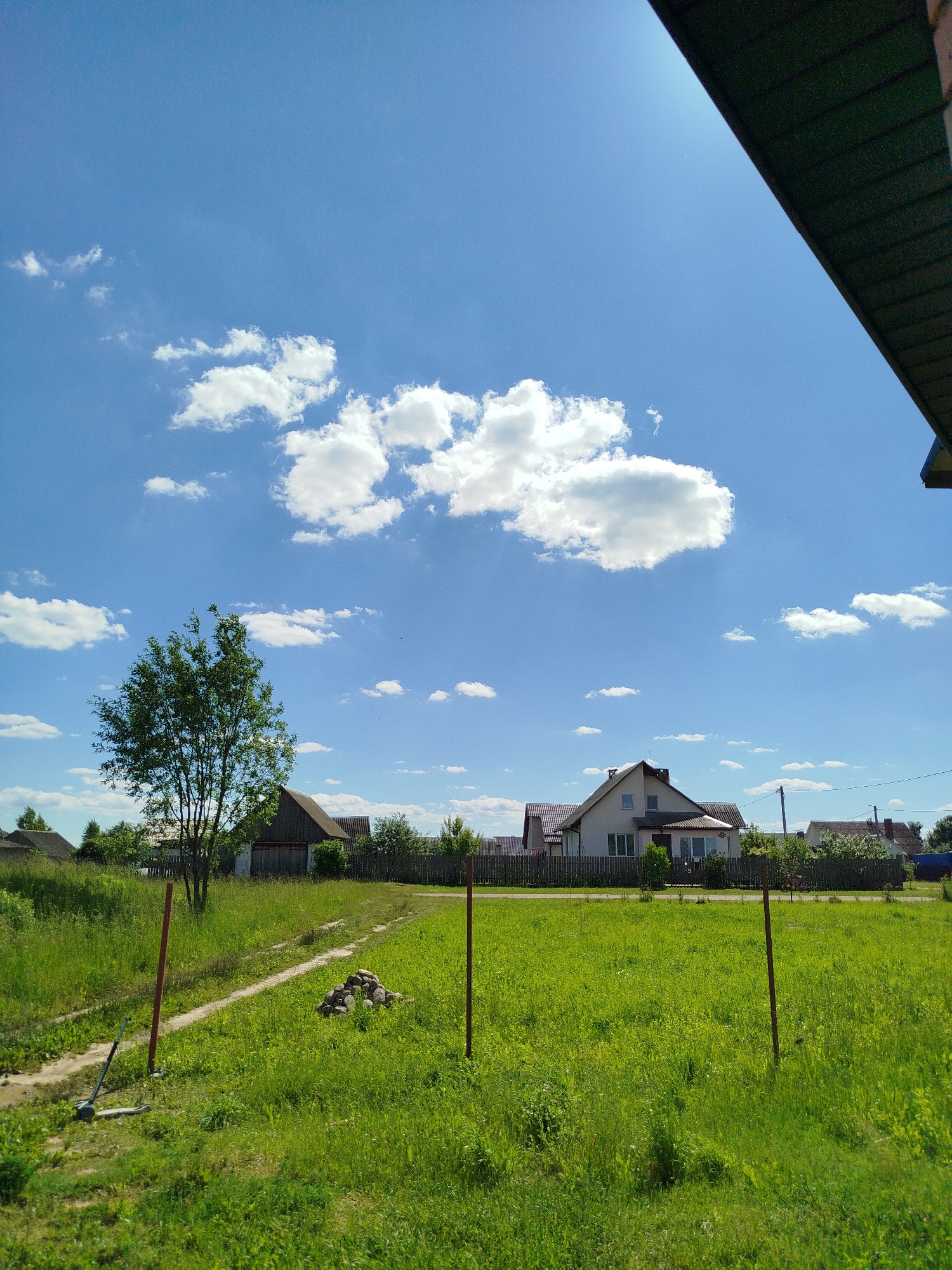 This image depicts a rural landscape under a bright blue sky dotted with fluffy white clouds. In the foreground, a patch of tall, green grass is interspersed with a dirt path. Two reddish-brown posts mark a boundary, and a small pile of stones sits on the grass. A grey kick scooter rests on the left side of the path. A lone, leafy green tree stands to the left of the frame, its branches reaching towards the sky.
In the middle ground, several houses with peaked roofs are visible behind a wooden fence. These houses appear to be residential buildings, with some featuring chimneys. Further in the background, more trees and vegetation line the horizon, suggesting a spread-out rural community. A utility pole with wires is also visible among the houses.
The overall scene suggests a sunny day, likely in the late morning or early afternoon, given the bright sunlight casting shadows. The setting is a peaceful, rural area, possibly in Belarus, given the provided location context. There is no text visible in the image. The atmosphere is tranquil and ordinary, with no specific activity or event being captured, other than a general view of a residential rural area on a pleasant day.