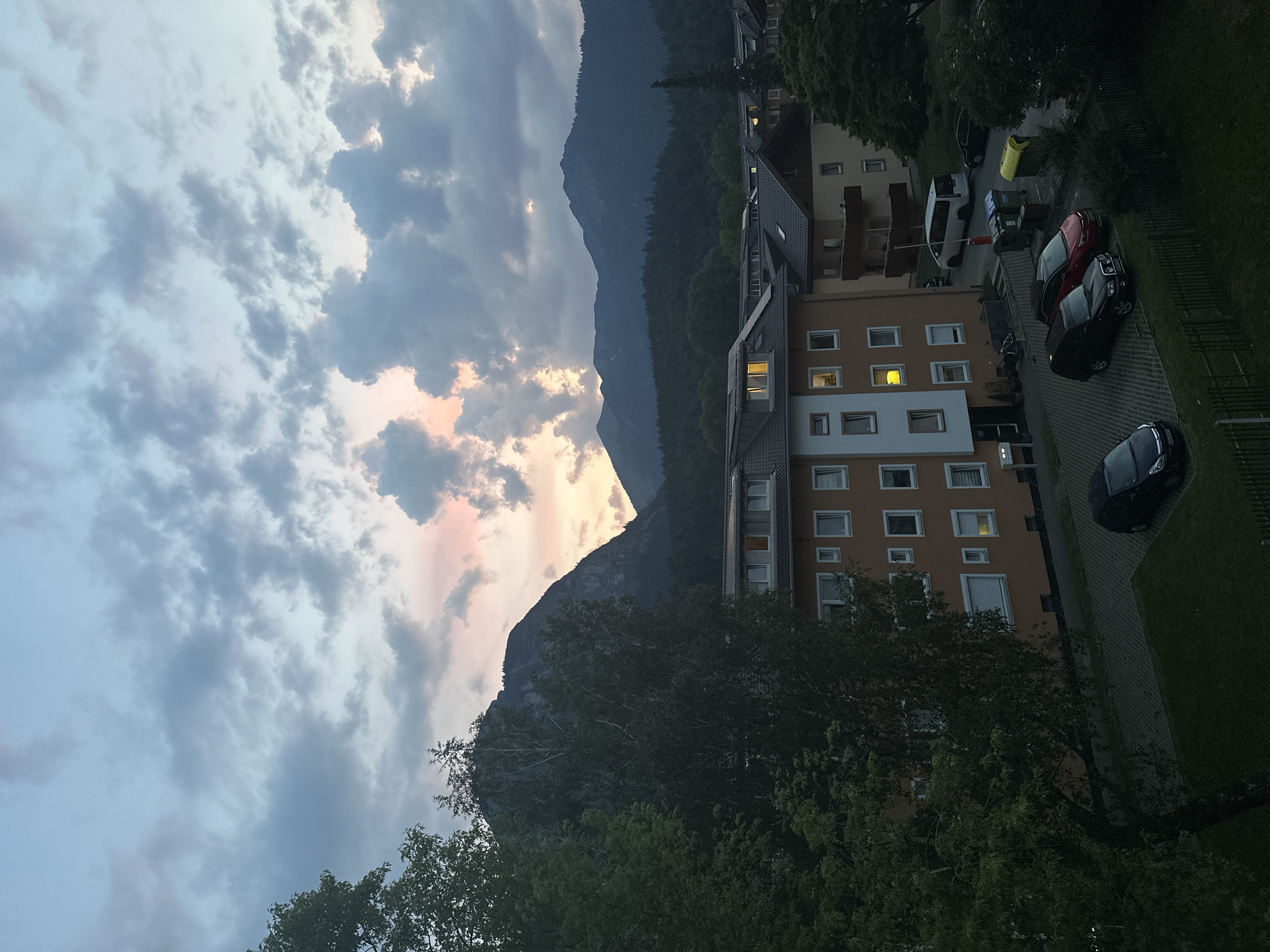 This is an outdoor, eye-level, medium shot of a residential area in Garmisch-Partenkirchen, Germany, at dusk. The foreground is dominated by the lush green foliage of a large tree on the left and the dark shadow of a fence. Beyond the tree, several residential buildings are visible. The closest building is orange with white-trimmed windows, some of which are illuminated. Other buildings, with dark roofs and lighter facades, are also present. In front of the buildings, a paved parking area holds several cars, including a black SUV, a red sedan, and a white van. Garbage bins are also visible in the parking area. The midground features a dense forest leading up to the imposing mountains that form the backdrop. The sky above is filled with dramatic clouds, tinged with hues of pink and orange from the setting sun, contrasting with the cooler blues and grays of the overcast sky. The overall atmosphere is tranquil and serene.
