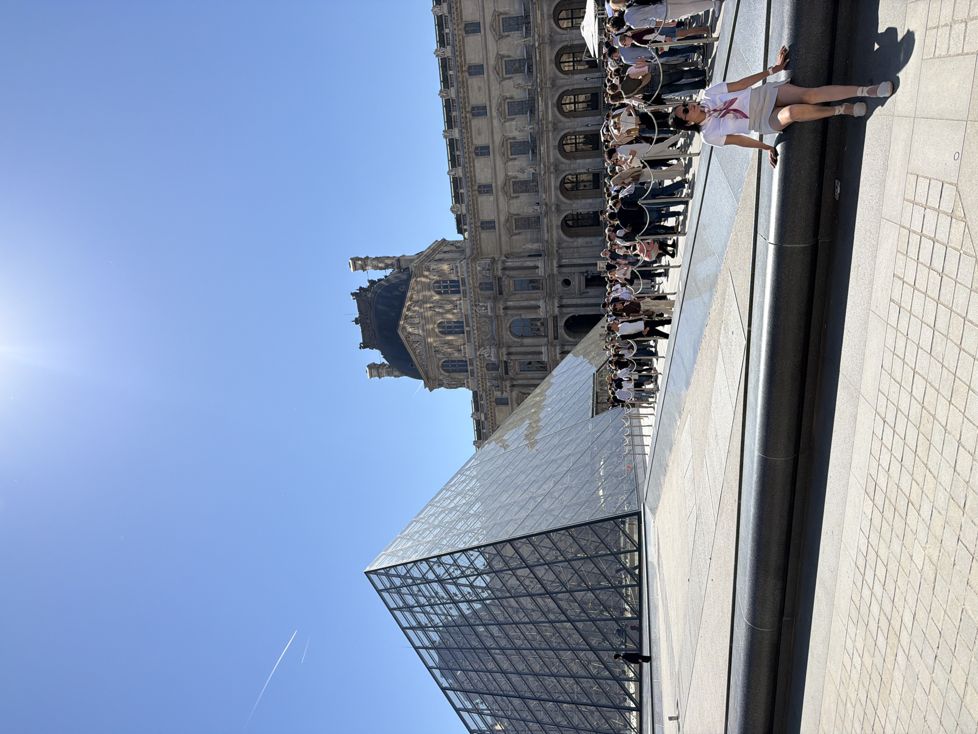 The image shows the Louvre Museum in Paris, France on a sunny day. The glass pyramid is prominent in the foreground. Behind it stands the Louvre Palace, with its ornate facade. A line of people is waiting to enter the museum. A woman lies on a ledge at the edge of the frame.