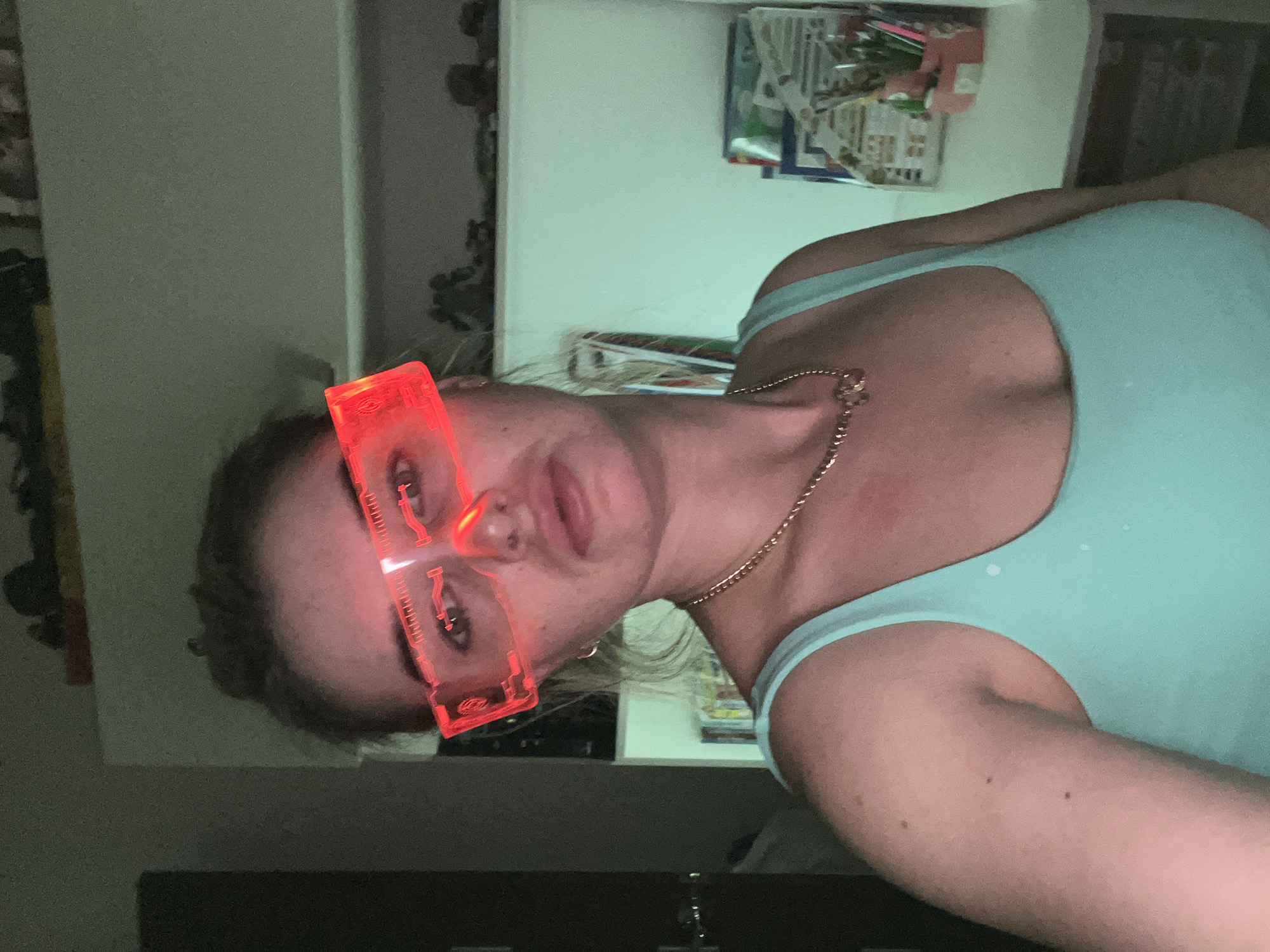A young woman with red, futuristic-looking eyewear is positioned in front of what appears to be a shelf or cabinet. She is wearing a light blue tank top and a gold chain necklace. The shelf contains various items. The room is dimly lit. There is no definitive evidence in the image to verify the location as Chaikovsky, Russia.