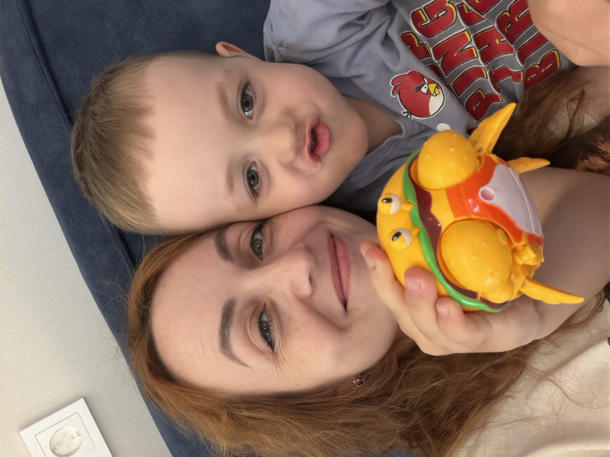A woman and a young child are lying down next to each other. The child is wearing a gray shirt with an Angry Birds design. The woman is holding a yellow and orange plastic toy. They are lying on a blue surface. A light switch is visible in the lower left corner. The city is Chaikovsky, Russia.