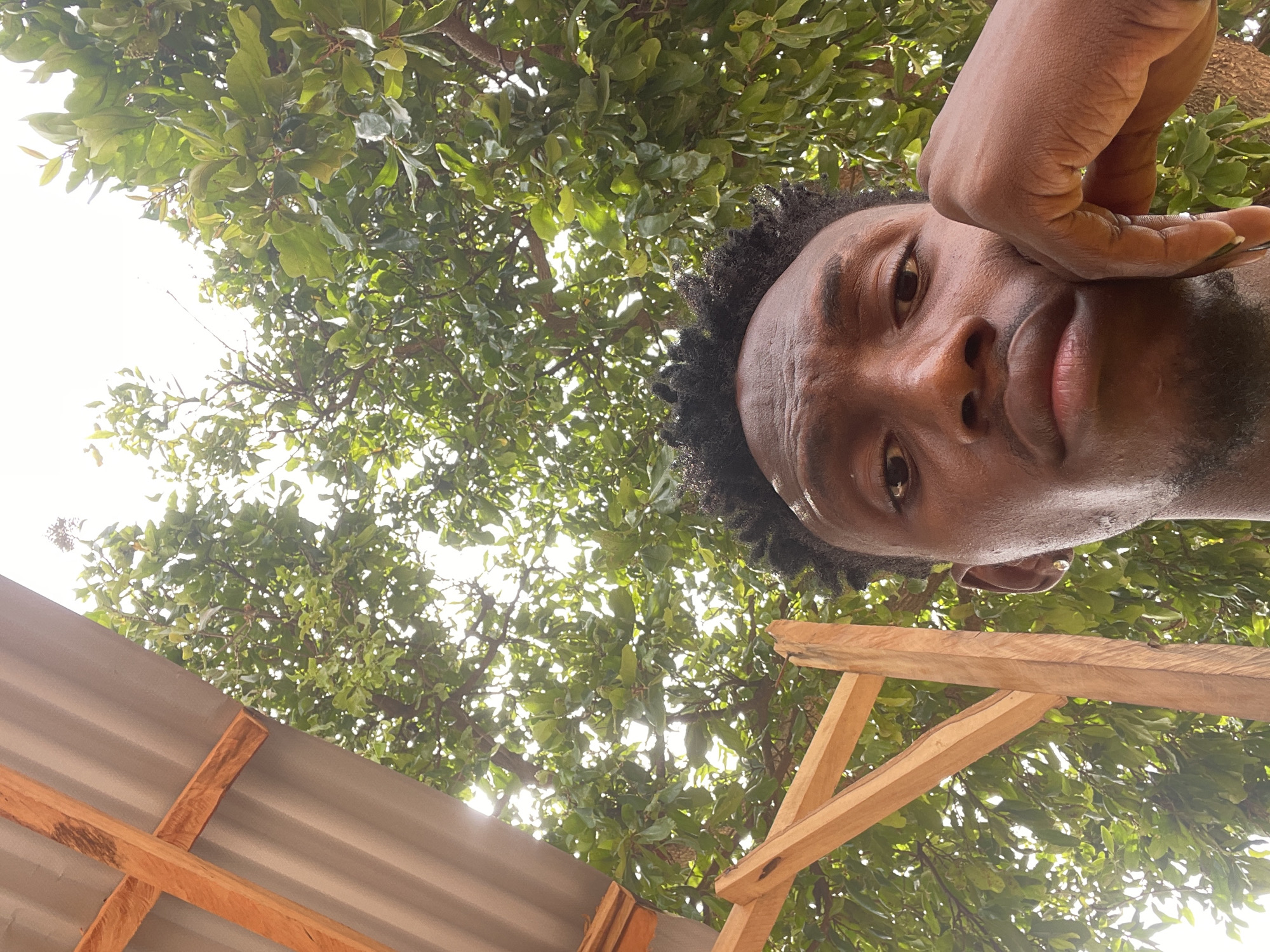A low-angle shot shows a person with dark skin, short curly hair, and a trimmed beard, positioned with their face close to the camera. Their hand is raised near their cheek. Above them is a canopy of green foliage, suggesting a tree. Below, sections of a wooden frame and corrugated metal are visible, possibly a roof structure. The location is indicated as Lokoja, Nigeria, but there are no specific city-defining landmarks visible.
