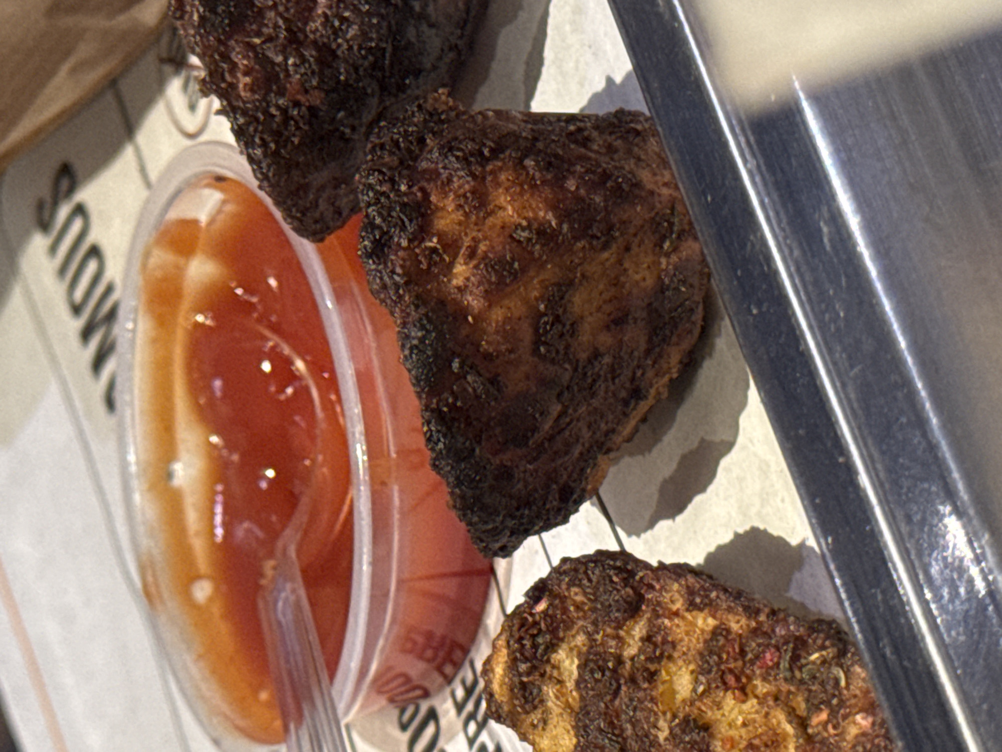 The image displays a close-up, high-angle shot of several pieces of darkly cooked food, likely grilled or fried chicken, alongside a small cup of dipping sauce. The food items have a visibly crispy, textured, and somewhat charred exterior, varying in shades of dark brown and black, with some lighter golden-brown areas showing through.
To the left, a small, clear plastic cup is filled with a reddish-orange, somewhat viscous sauce. A clear plastic spoon is partially submerged in the sauce, resting against the rim of the cup.
The food and sauce container are placed on what appears to be a light-colored, possibly paper or cardboard, liner or packaging. Partial text is visible on this surface, including the letters "SMOUS" (likely part of "FAMOUS") oriented diagonally, and further down, "BBS" and "00%".
On the far right of the image, a sleek, reflective metallic surface is visible, running vertically. This could be part of a serving tray, a counter, or a food warmer, reflecting light. Shadows are cast by the food pieces on the light background, suggesting a light source from above and to the left. The overall impression is of a casual dining or take-out meal, with the food prepared for consumption.