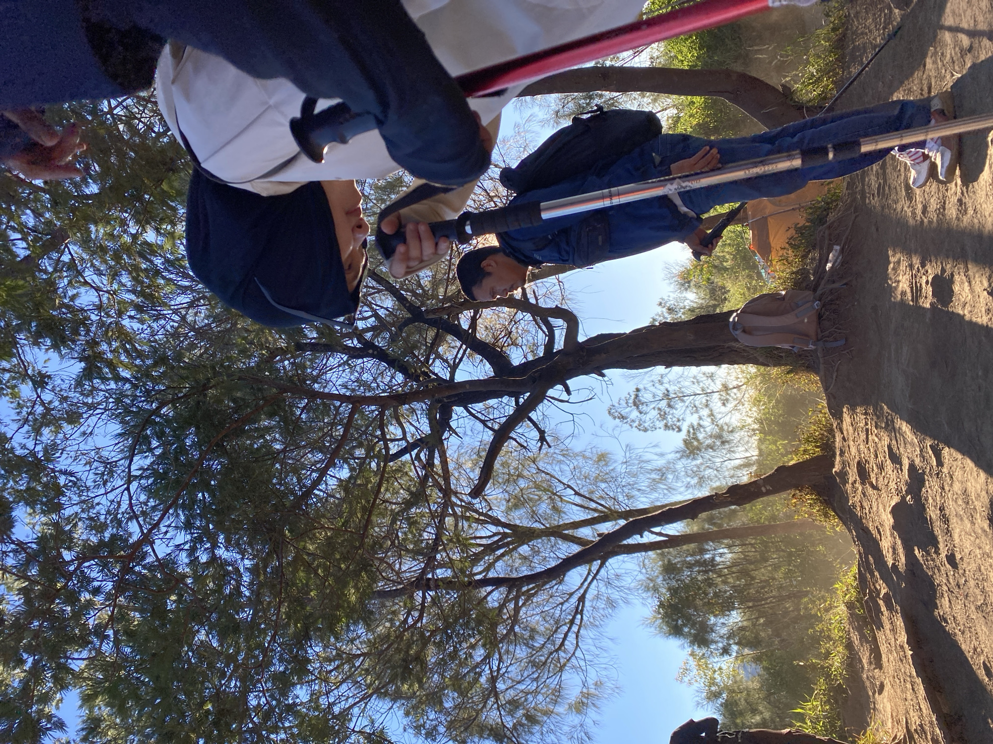 The image depicts a group of people on a dirt path with sparse trees and vegetation, characteristic of a high-altitude or volcanic landscape. The sun is shining brightly, casting long shadows on the ground. In the foreground, a person wearing a blue jacket and jeans is holding two trekking poles. Another person, wearing a white jacket and a black headscarf, is partially visible in the upper right, also holding a trekking pole. A backpack is visible on the ground near a tent in the background. The overall impression is of an outdoor activity, possibly hiking or trekking. The location is identified as Gondosuli, Indonesia, which is known for its volcanic landscapes and natural beauty.