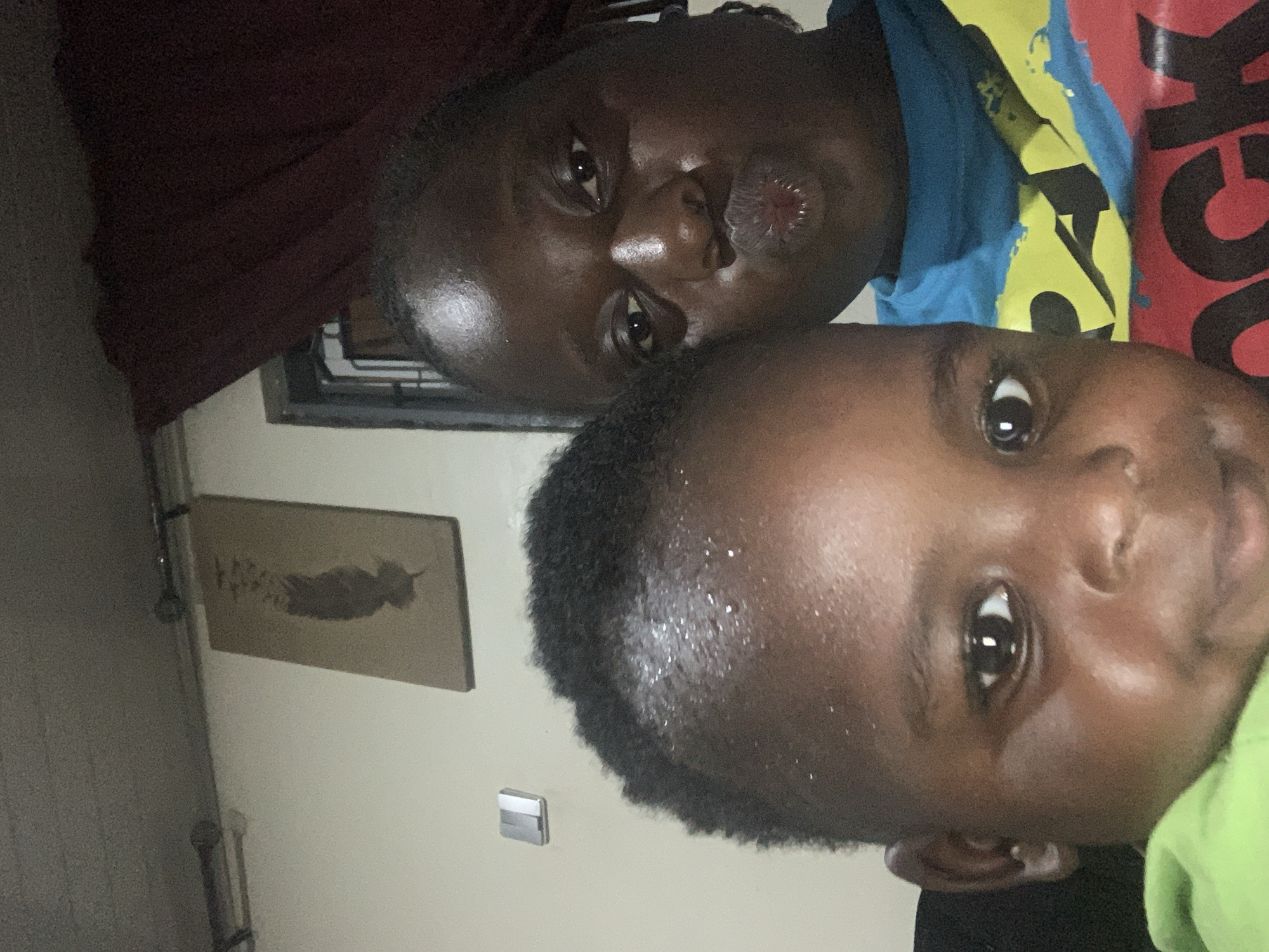The media file is a close-up, slightly over-the-shoulder selfie taken indoors. The subjects are two individuals, likely an adult and a child, with dark skin tones.
The adult is in the foreground and left side of the image, with their face turned towards the camera and puckered lips. They are wearing a dark maroon garment that covers part of their shoulder and arm. Behind them, a portion of the child's face is visible, with a wide open eye and part of their forehead and hair. The child is wearing a bright green garment on their right side. The background is blurred, but it appears to be a domestic setting. A framed picture of what looks like a silhouetted animal or person is hanging on the wall to the left. There is also a small, silver electrical outlet cover visible on the wall. The lighting suggests it is daytime, but the exact time and weather are not discernible. The overall mood appears casual and intimate, typical of a selfie.
There is some text visible on a colorful t-shirt worn by the adult. Part of the text reads "CK" in large black letters on a red background, and there is also a yellow and blue design with black shapes.