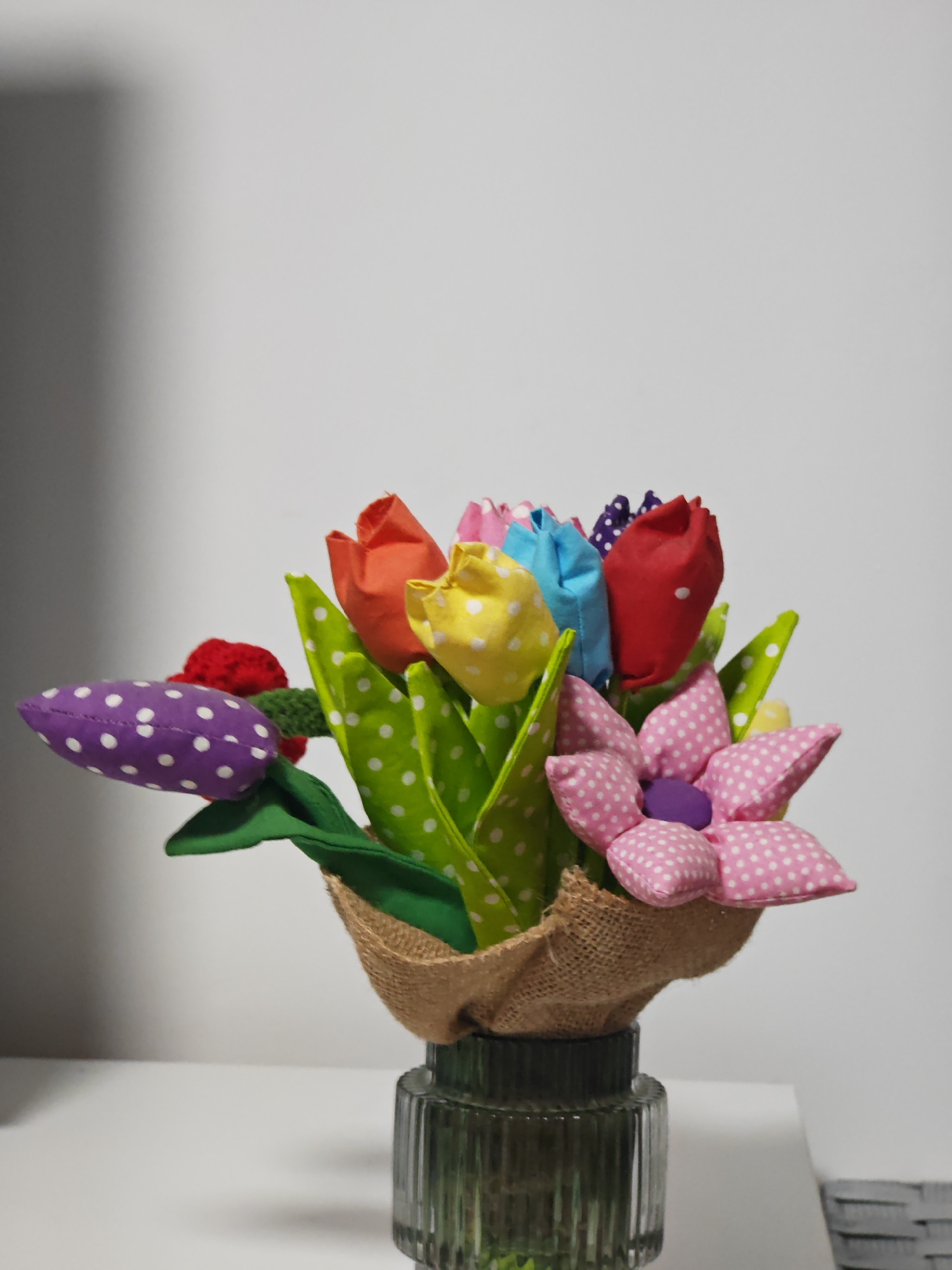 A static, eye-level photograph, taken in Shumen, Bulgaria, displays a bouquet of artificial fabric flowers situated on a white surface against a plain light grey wall. The bouquet includes multiple fabric flowers: several tulip-shaped blooms in red, yellow, light blue, and purple, each adorned with white polka dots, alongside bright green polka-dotted leaves. A prominent pink fabric flower with white polka dots and a purple center is also visible, accompanied by a small red knitted flower. The base of the bouquet is wrapped in light brown burlap and contained within a transparent, dark green, vertically ribbed glass vase. The foreground is a white, flat surface, while the background is an undifferentiated light wall exhibiting a soft shadow in the upper left. No people, actions, or interactions are depicted.