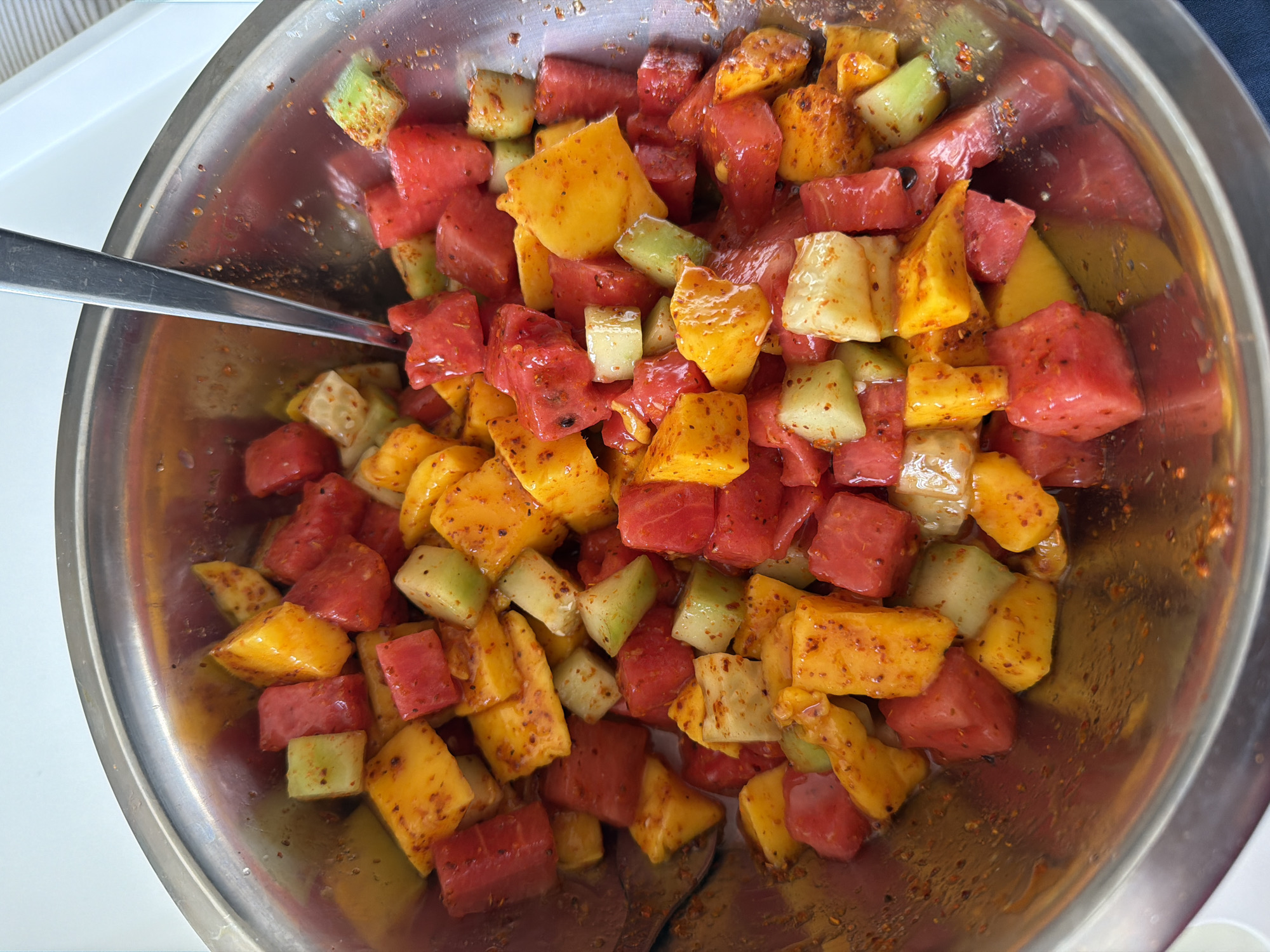 A top-down, close-up shot shows a vibrant fruit mix in a large, round, metallic mixing bowl, likely made of stainless steel. The bowl is filled with generously diced pieces of fruit, uniformly cut into cubes.
The fruit assortment includes bright red cubes, characteristic of watermelon, and yellow-orange pieces that appear to be mango or possibly pineapple. Scattered throughout are lighter green or off-white cubes, consistent with cucumber or jicama. All the fruit pieces are thoroughly coated with a reddish-brown, speckled seasoning, which strongly resembles chili powder or a chili-lime blend.
A silver-colored spoon is positioned within the bowl, resting among the fruit, suggesting the mixture is either being stirred or is ready to be eaten. A small amount of liquid, likely fruit juices combined with the seasoning, is visible at the bottom of the bowl. The metallic surface of the bowl reflects light, and some minor smudges are discernible. The background is out of focus, revealing clean, light-colored surfaces, indicative of a kitchen or dining setting. The overall impression is of a refreshing and flavorful fruit preparation. There is no visible text or people in the image.