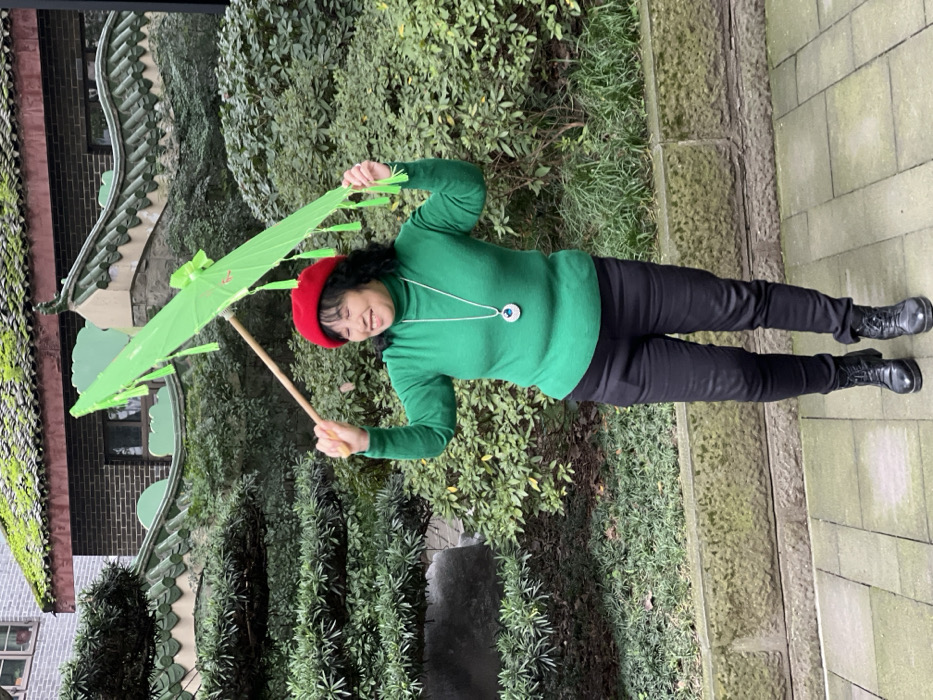 A woman in a green sweater and red beret poses outdoors with a green parasol. She wears black pants and boots. The background includes a traditional building with a green tiled roof and a courtyard with greenery. The woman smiles and holds the parasol above her head.