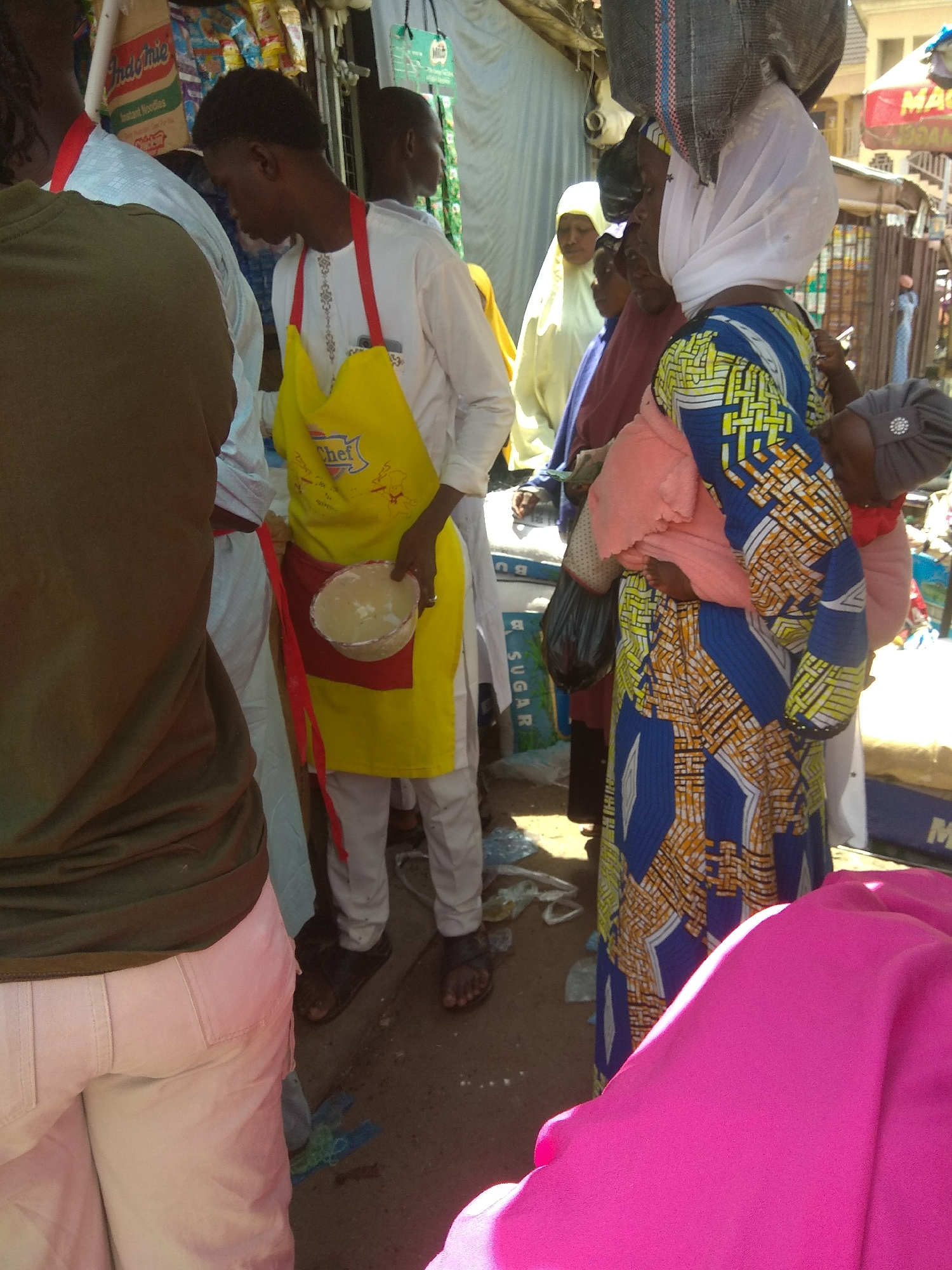 This image shows a bustling outdoor market scene in Jalingo, Nigeria. The focal point is a young man wearing a white shirt and a bright yellow apron with "Chef" printed on it. He holds a round, shallow bowl containing a light-colored substance, possibly food or ingredients. He is standing amidst a crowd of people.
To his left, another individual, partially visible from the back, wears a dark green t-shirt and light-colored pants. To his right, a woman in a vibrant blue, yellow, and orange patterned dress and a white headscarf carries a baby wrapped in a pink cloth on her back. Other people are visible in the background, some wearing headscarves and traditional attire, suggesting a diverse group.
The setting appears to be a market stall with various goods displayed, including bags of what appears to be "SUGAR." In the background, there are awnings and structures indicating a lively commercial area. The lighting suggests it is daytime, likely a sunny day due to the shadows. The overall atmosphere seems busy and active, typical of a marketplace. The people's expressions are not clearly discernible, but the scene conveys a sense of daily commerce and community interaction.