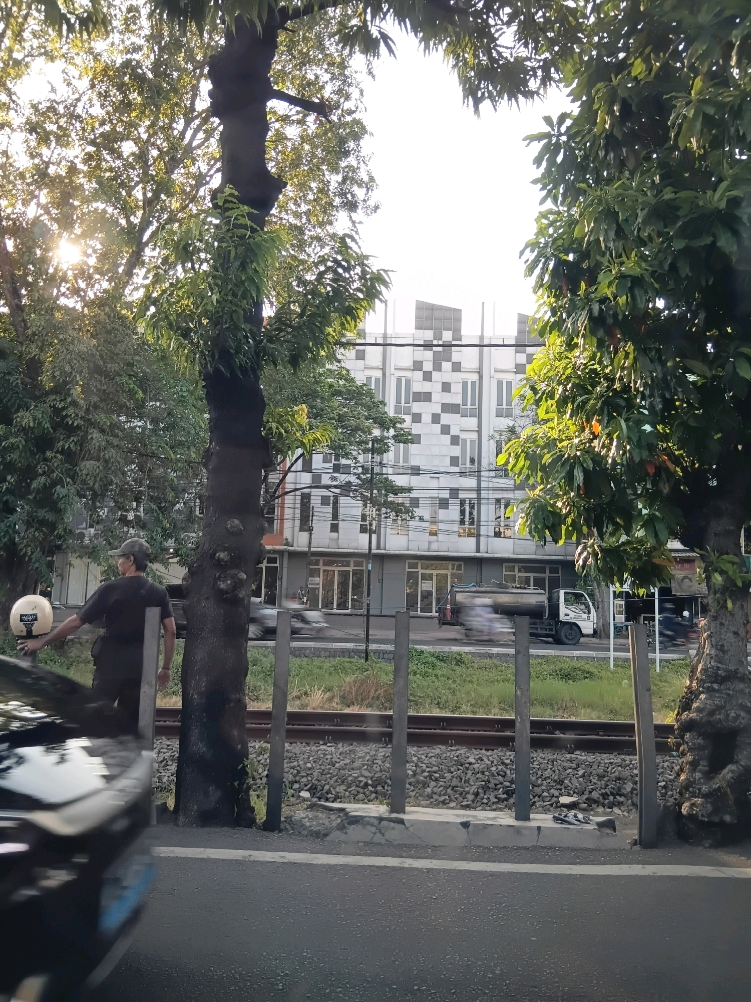 In Surabaya, Indonesia, a man in a dark t-shirt and dark pants, wearing a camouflage baseball cap, stands near a tree, holding a white helmet with a black strap in his left hand. He is looking to his left, towards a blurred black vehicle in the foreground. Behind him, a modern building with a white facade and a geometric pattern of grey squares and rectangles rises against a bright sky. A railway track with gravel ballast runs horizontally across the mid-ground, bordered by wooden posts and a concrete curb. A white truck with a tank is visible in motion on the road behind the railway tracks, along with other blurred vehicles and pedestrians. Lush green trees frame the scene, with the sun's rays filtering through their leaves, creating dappled light. The foreground features a section of dark asphalt road.