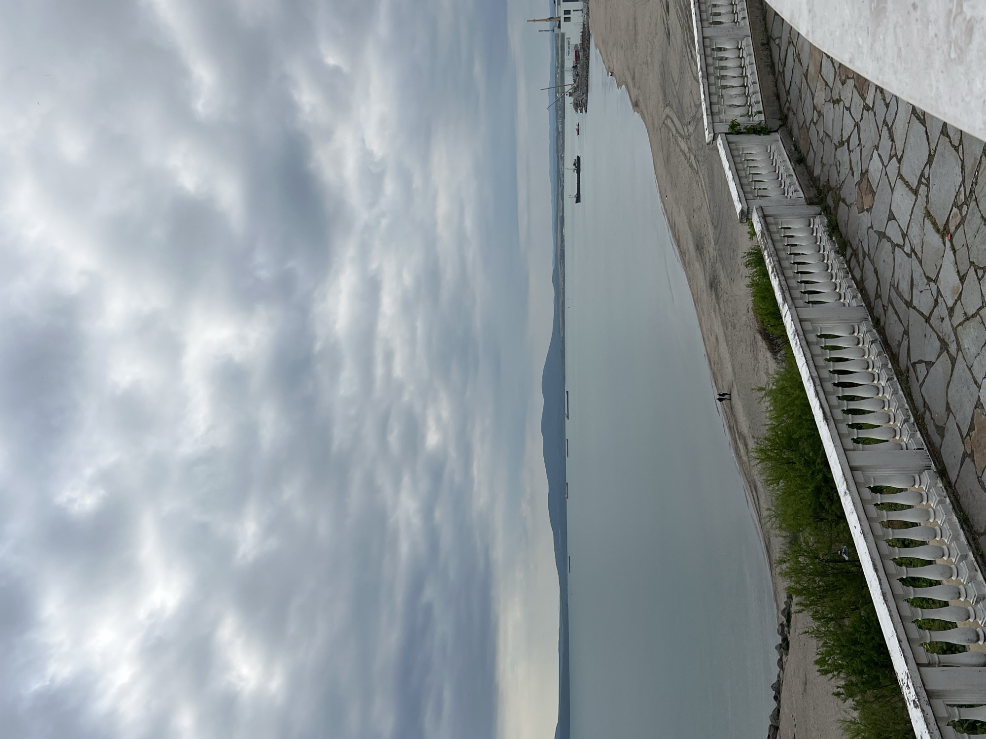 The image shows a view of the Black Sea from what appears to be the Sea Garden in Burgas, Bulgaria. The sky is heavily overcast with dense clouds. A body of water stretches into the distance, likely the Burgas Bay. In the far background, there are hints of land and some industrial port infrastructure including cranes. A ship or barge can also be seen in the water.
On the right side of the image, a stone walkway with a decorative white balustrade runs along the edge. Green shrubbery is visible growing along the base of the balustrade. The ground surface is made of irregularly shaped stone slabs.