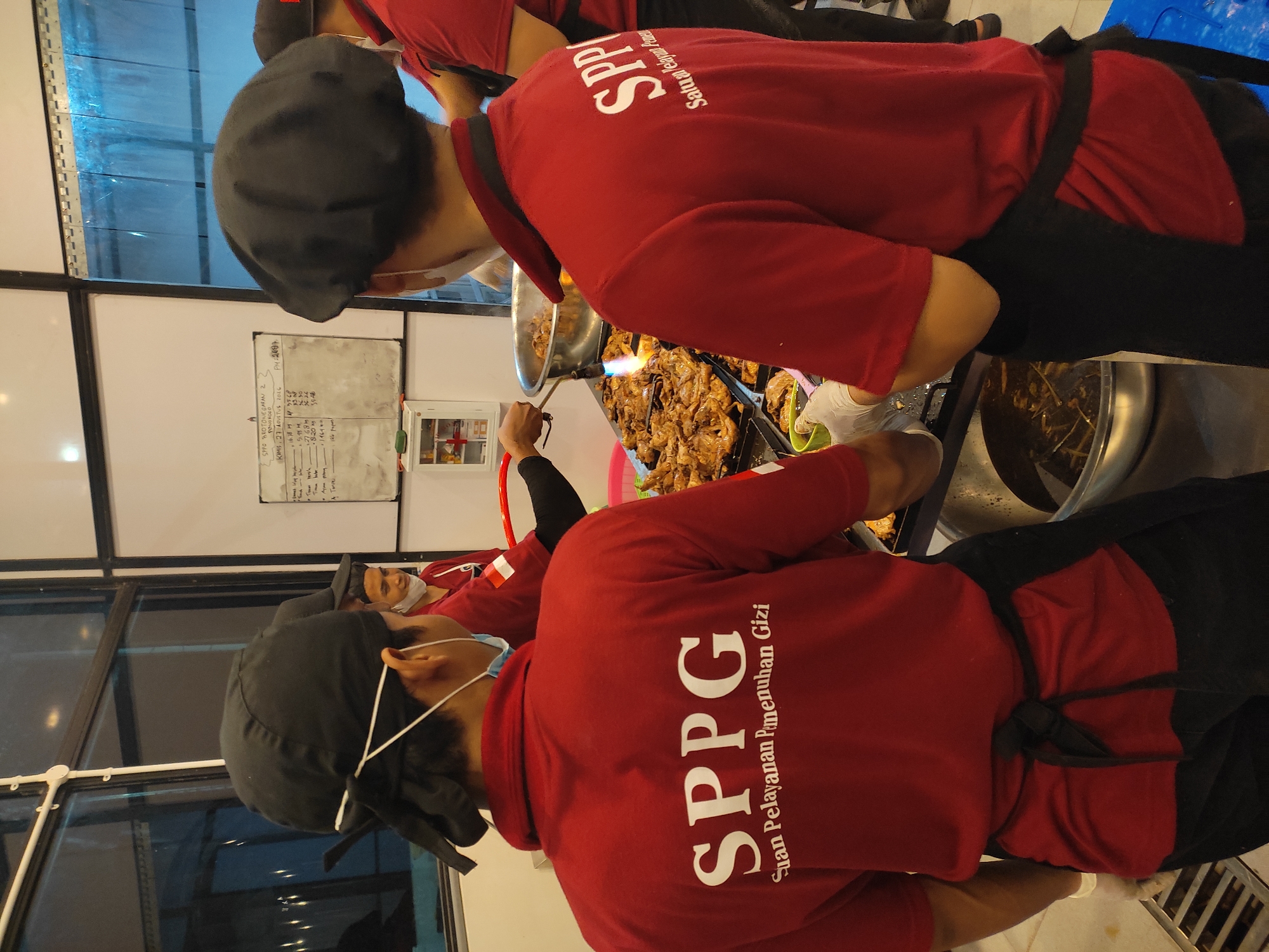 In Ponorogo, Indonesia, a culinary preparation is underway, likely within a food service establishment or event. Two individuals, presumed to be staff, are actively engaged in cooking. Both are wearing red shirts with the prominent white lettering "SPPG" and the text "Satuan Pelayanan Pemenuhan Gizi" below it. They are also wearing black head coverings and one is wearing a black apron tied around their waist.
The scene is focused on a long table laden with numerous cooked chicken pieces. In the background, a large metal pot or basin is visible, containing some liquid and what appears to be more food items. One of the individuals is holding a torch-like device, directing a flame towards the chicken, suggesting a grilling or caramelizing process. Another individual is holding a green bowl and wearing white disposable gloves, possibly preparing to serve or plate the food. A whiteboard with handwritten text and numbers is mounted on the wall in the background, which may contain inventory, orders, or schedules. The overall environment appears to be an indoor space with industrial-style lighting and what might be a plastic curtain partition in the far background.