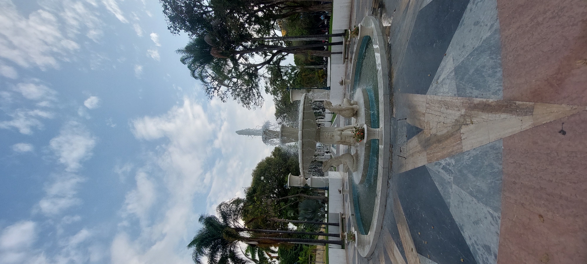 The image depicts a fountain in a plaza in Caracas, Venezuela. The fountain is multi-tiered and features statues of figures at each level. Water is actively flowing from the fountain. The plaza is paved with a geometric pattern of dark and light colored stones. The sky is blue with scattered clouds. Trees surround the plaza.