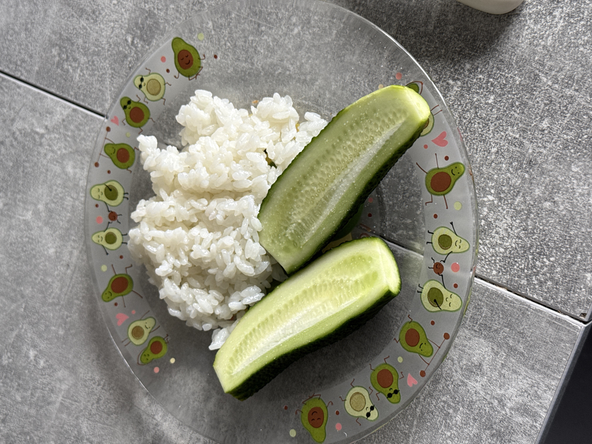 A close-up overhead shot captures a clear glass plate adorned with cartoon avocados on a gray, textured surface. The plate contains a mound of white rice and two halved cucumbers, with their white flesh and seeds visible. The gray surface appears to be tile or stone. The scene is indoors, likely in a kitchen or dining area, but its specific location in Lyubertsy, Russia, cannot be determined from the visual information provided.