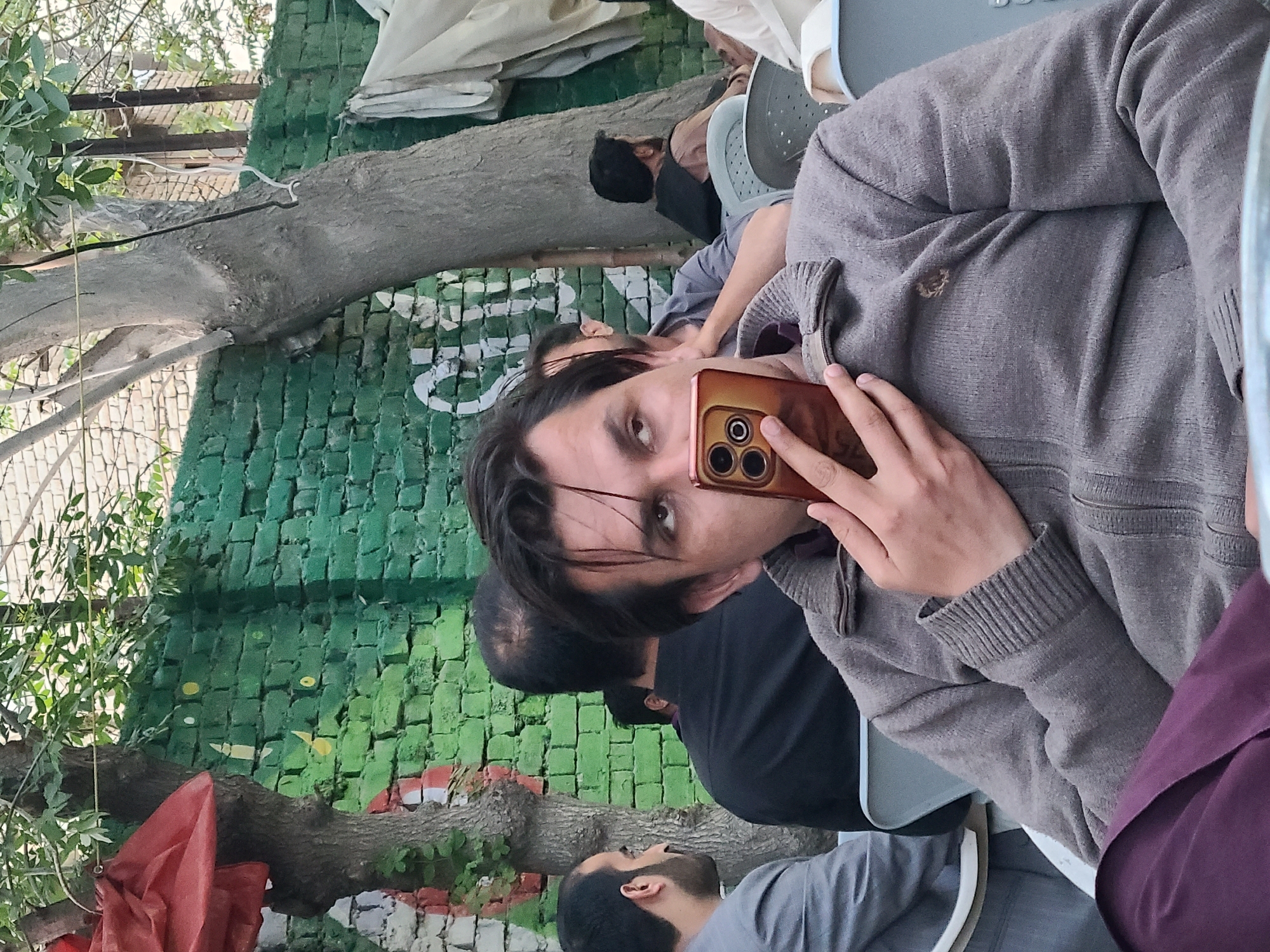 The image depicts a group of people gathered in an outdoor setting in Quetta City Tehsil, Pakistan. A man is holding a smartphone up to his face as if taking a selfie. He is wearing a gray sweater. Other individuals are sitting nearby. The background features a green brick wall with graffiti. There are trees present.