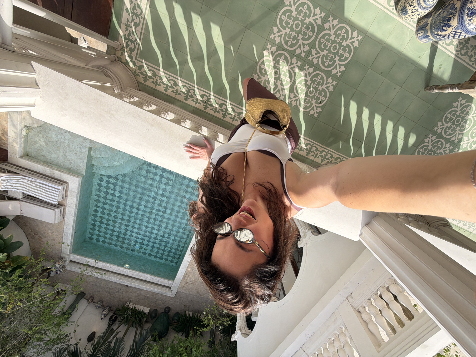 This high-angle selfie captures a young woman looking down towards the camera, her head tilted, revealing a bright smile. She has long, wavy dark hair and wears reflective sunglasses, a white sleeveless top, and a brown garment (possibly shorts or a skirt). A delicate gold chain necklace and a small, textured golden handbag with a chain strap complete her look.
The scene below her features a distinctive light green tiled floor adorned with an intricate white geometric and floral pattern. Strong, wavy shadows from an unseen structure are cast across these tiles, indicating bright, sunny weather, likely midday or early afternoon. White, classical architectural elements, including a decorative balustrade and carved detailing, form the foreground, suggesting a balcony or upper terrace.
Further into the scene, a rectangular swimming pool with a light blue and white checkerboard tile pattern at its bottom is visible. Poolside lounge chairs with striped cushions are positioned next to the pool. The surrounding area is lush with green foliage, trees, and decorative elements such as pots and sculptures, contributing to a tropical or resort-like atmosphere. Two large, blue and white patterned ceramic vases are also present on the patterned floor. The setting appears relaxed and leisurely, consistent with a vacation in North Kuta, Indonesia. No discernible text is visible.