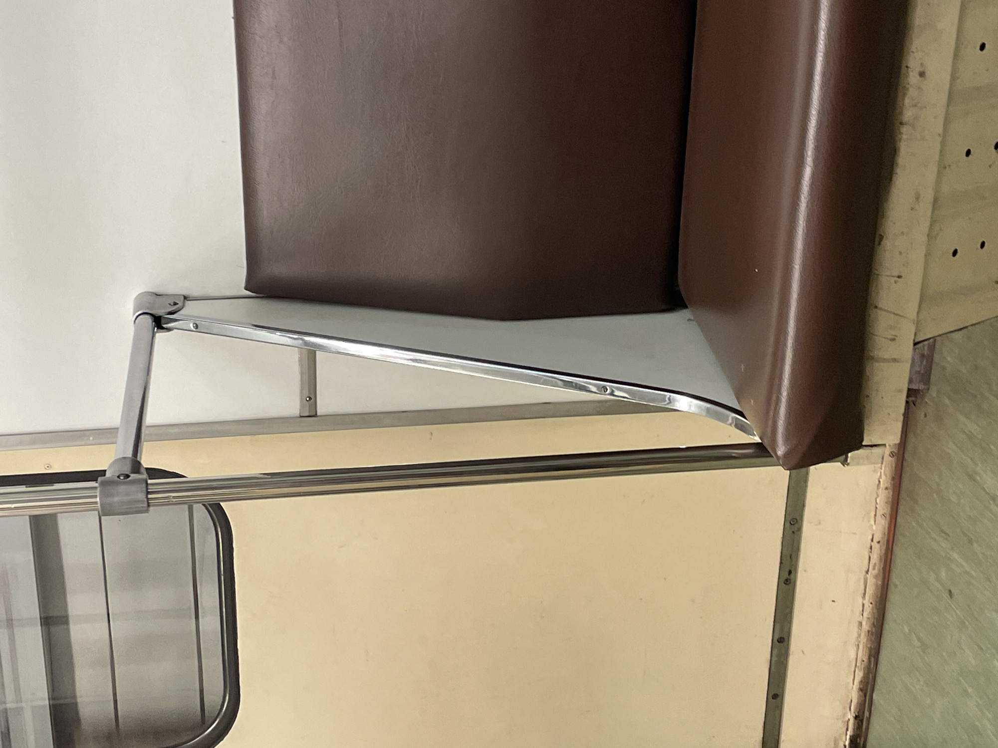 The image shows a close-up view of a brown vinyl seat and a metal handrail inside a train car. The seat appears to be worn, with some creases and a slightly scuffed texture. The handrail is shiny and metallic, attached to a white wall. To the left of the seat is a cream-colored door with a large, rectangular window. The floor is a light green linoleum with a subtle speckled pattern. The overall impression is of a utilitarian interior, likely a subway or commuter train. The specific location in Moscow, Russia, cannot be determined from the visual information provided.