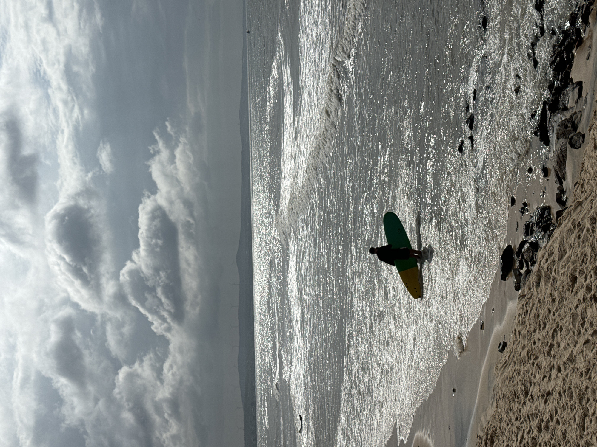 A solitary individual stands in the shallow water at the edge of a beach, holding a colorful surfboard. The surfboard has a yellow and green design and is partially submerged in the water. The person, silhouetted against the bright, choppy sea, is wearing dark clothing. The ocean exhibits a high level of reflected light, creating a shimmering effect on its surface, with visible waves rolling towards the shore. The beach in the foreground consists of sand and scattered rocks. In the distance, a hazy silhouette of a coastline with mountains and wind turbines is visible beneath a heavily clouded sky. The overall atmosphere is overcast and possibly windy. The scene does not depict any specific landmarks or features that would definitively place it within Phan Thiết, Vietnam.