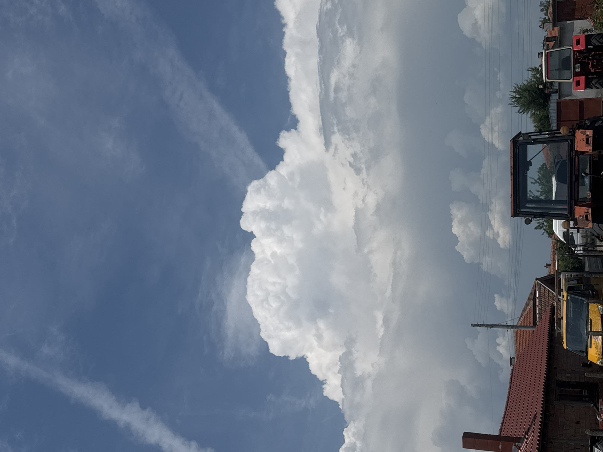 This image depicts a daytime scene in Sarnevo, Bulgaria, characterized by a dynamic sky and a ground level featuring residential structures and agricultural vehicles. The upper portion of the sky is a clear, bright blue, traversed diagonally by two distinct, thin white contrails from aircraft. Below this, a massive formation of bright white cumulus clouds dominates the mid-sky, exhibiting significant vertical development. Towards the horizon, these clouds become denser and appear slightly darker grey.
At ground level, on the left, a building with a red-tiled roof and a brick chimney is partially visible. Adjacent to it, a yellow utility van or small truck, equipped with a roof rack holding a ladder, is parked. In the central part of the frame, a tall wooden utility pole supports multiple horizontal power lines that stretch across the mid-ground. To the right of the yellow van, an orange-red tractor is prominently positioned, facing the viewer. Further to the right and slightly behind, a second, smaller tractor, primarily red with white accents, is also parked facing the viewer. Additional red-tiled roofs, various green trees, and other indistinct structures are visible in the background, interspersed between the utility lines and vehicles. No people are present in the image.