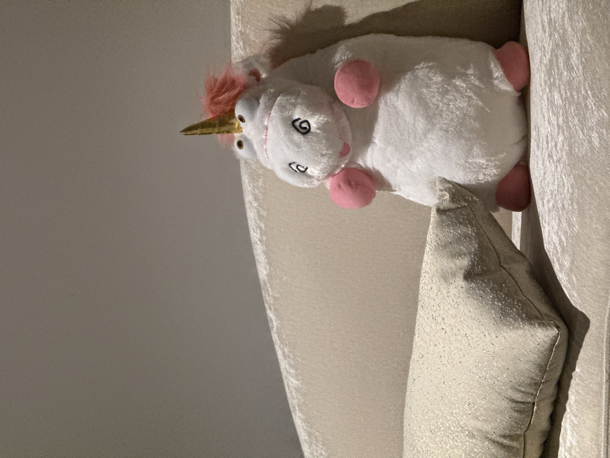 The image displays a white unicorn plush toy lying on a light-colored, possibly velvet, surface, with a textured, light-colored pillow beside it. The unicorn features a golden horn, a pink fluffy mane, pink hooves, and an embroidered face with large, oval eyes and a smiling mouth. Its body is plush and appears soft. Behind the plush toy and pillow, a plain grey wall is visible. The scene suggests an indoor setting, likely a bedroom or living space, in San Bernardino Tlaxcalancingo, Mexico. The lighting indicates an ambient indoor environment, with soft shadows. No specific activity is occurring other than the toy resting in place. There is no visible text in the image.