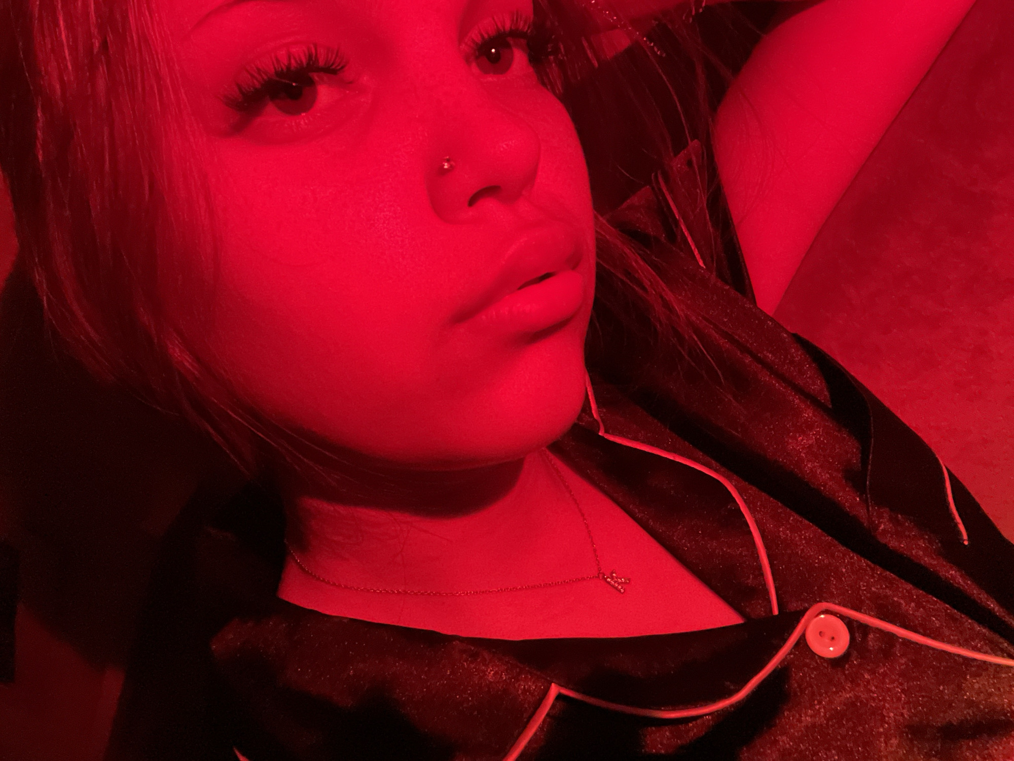 The image is a close-up, low-angle shot of a person illuminated by red light. The subject appears to be female, with long eyelashes and a nose piercing. She is wearing a dark-colored pajama top with a contrasting red trim and a single button. A delicate gold necklace with a small, possibly lettered, pendant hangs around her neck. The background is dark and out of focus, with the red light creating a strong, atmospheric glow. The image is cropped to focus on the subject's face and upper torso. The location is not discernible from the image.