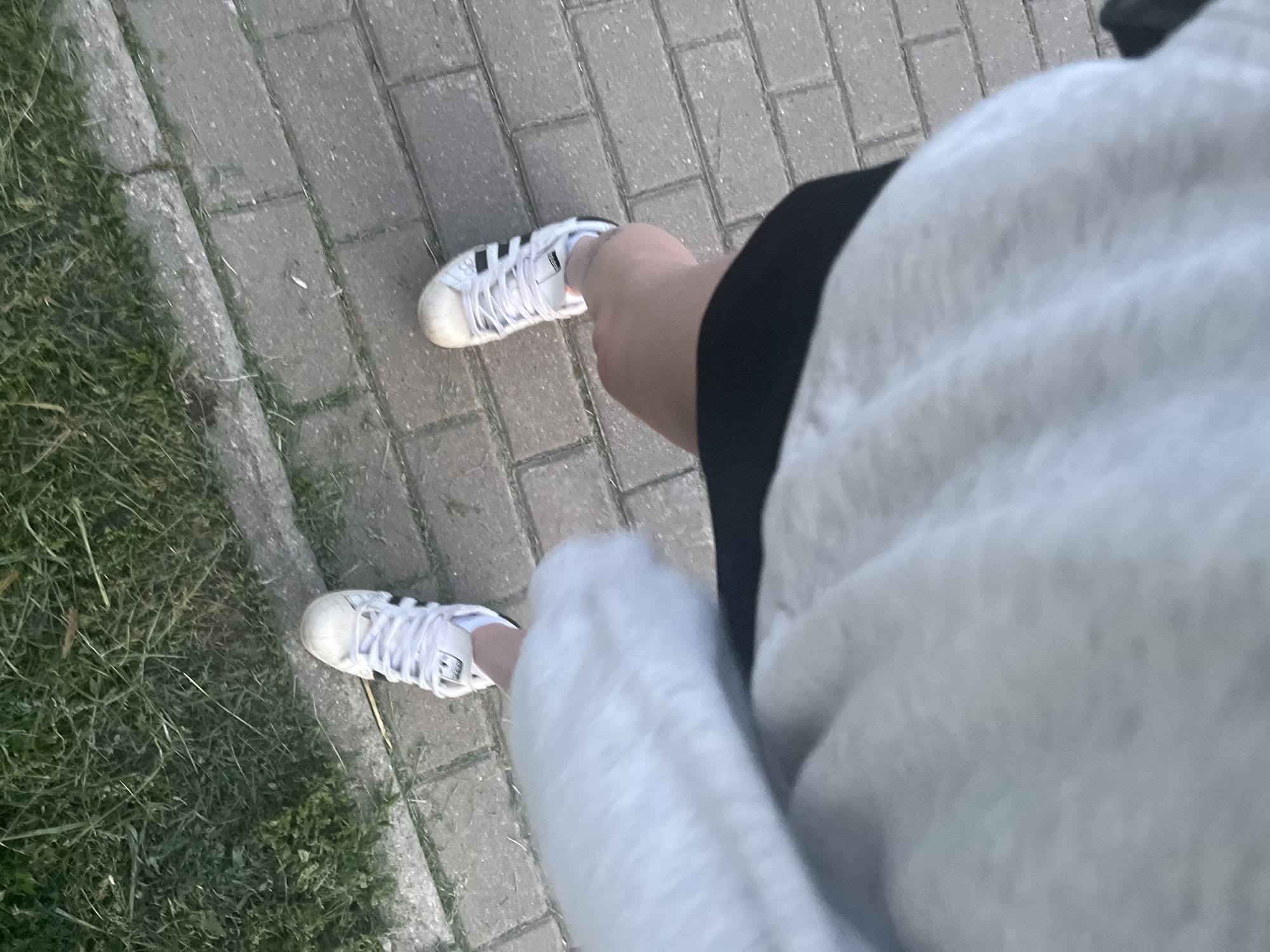 An overhead, first-person perspective photograph captures a person's legs and feet on a paved outdoor path in the city of Unknown, Unknown. The individual is wearing white Adidas Superstar sneakers with black stripes. Their left leg is partially obscured by light-colored, possibly white or light grey, fabric. A glimpse of dark fabric, likely shorts or a skirt, is visible between the light fabric and the legs. The paved path consists of rectangular grey pavers. To the left of the path, a strip of green grass with some weeds and dry blades is visible. The scene is illuminated by natural daylight.