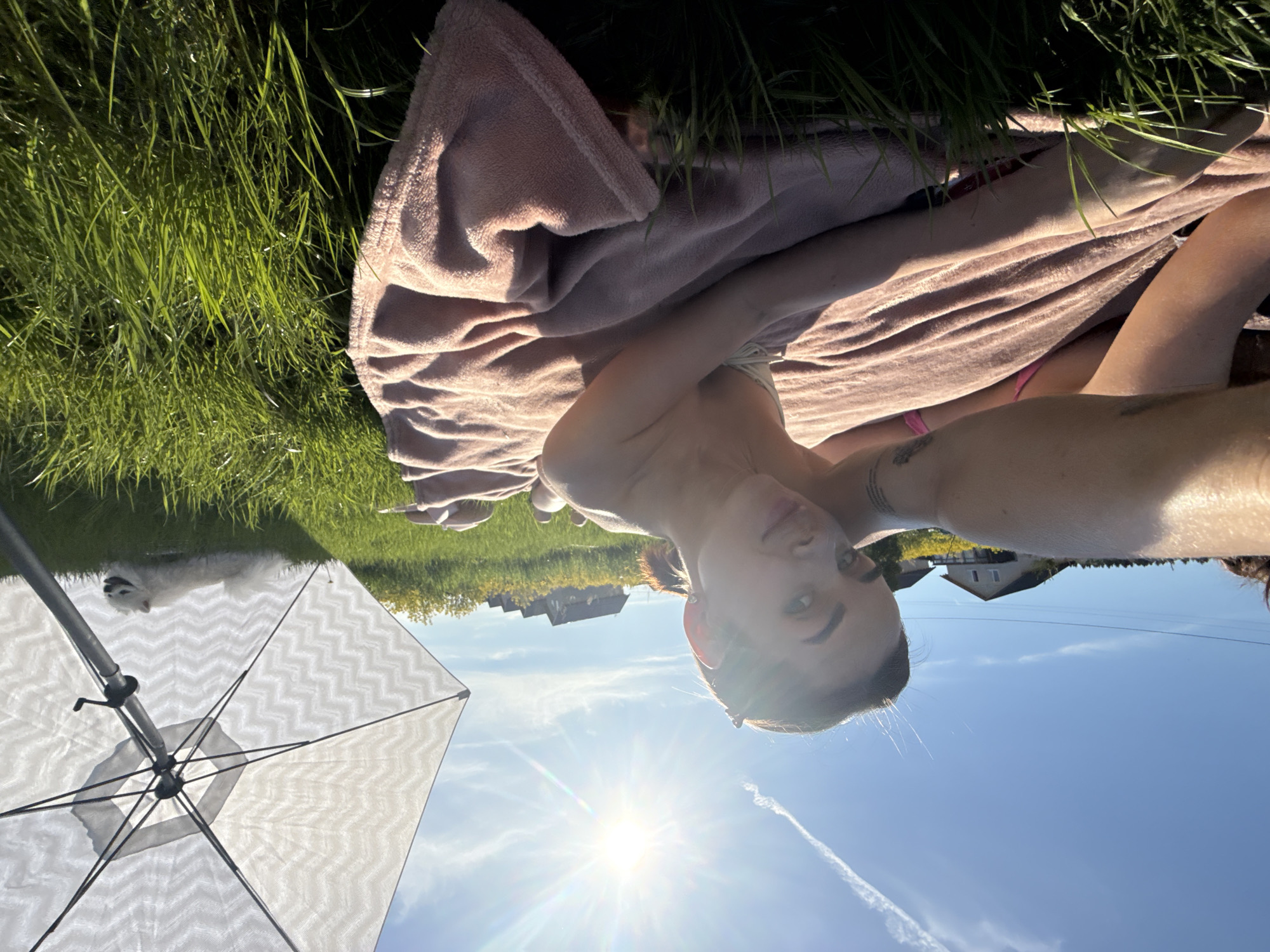 A woman is lying on a pink towel in the grass. A white dog is near her feet. An umbrella is to the left of the woman. The sun is shining brightly, with a contrail in the blue sky. In the background, there are houses, suggesting the location is in a residential area of Лобаново, Russia.
