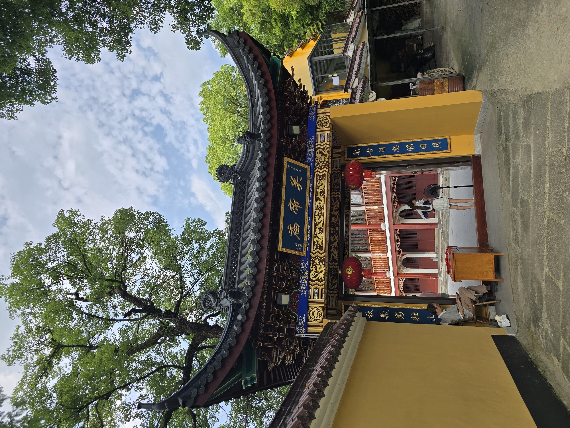 The image captures an entrance to a traditional Chinese courtyard, likely part of a temple or cultural site, situated in Xiaoshan District, China. The architectural style is characterized by a sweeping, ornate roof with dark tiles and intricate detailing, supported by colorful beams and columns. A prominent golden plaque with blue Chinese characters is affixed to the archway, above a large red lantern.
To the left of the entrance, a man in a grey shirt and beige pants sits on a wooden chair, looking at a mobile phone. Beside him is a wooden desk with a red cloth draped over it. To the right of the entrance, a young woman in a white dress stands near a pedestal fan, facing towards the courtyard.
The courtyard itself features a multi-story building with red balconies and windows, suggesting residential or administrative quarters within the complex. The ground is paved with stone, and the area is surrounded by lush green trees, indicating a natural or well-landscaped environment. The sky above is partly cloudy, with patches of blue visible. The overall impression is one of serenity and cultural heritage.