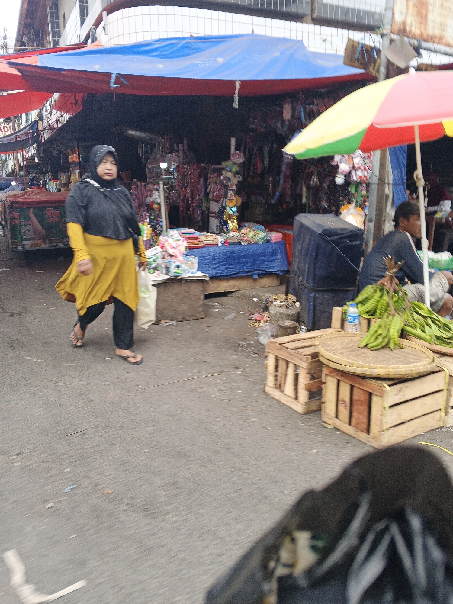 In Sukabumi, Indonesia, a woman in a black hijab and a yellow tunic walks across a street market. She carries a white plastic bag in her right hand and wears sandals. Behind her, various market stalls display goods under colorful tarps. A man is seated near a stall displaying green vegetables. Wooden crates and a woven mat are visible in the foreground. The scene is a busy marketplace with a mix of people and vendors.