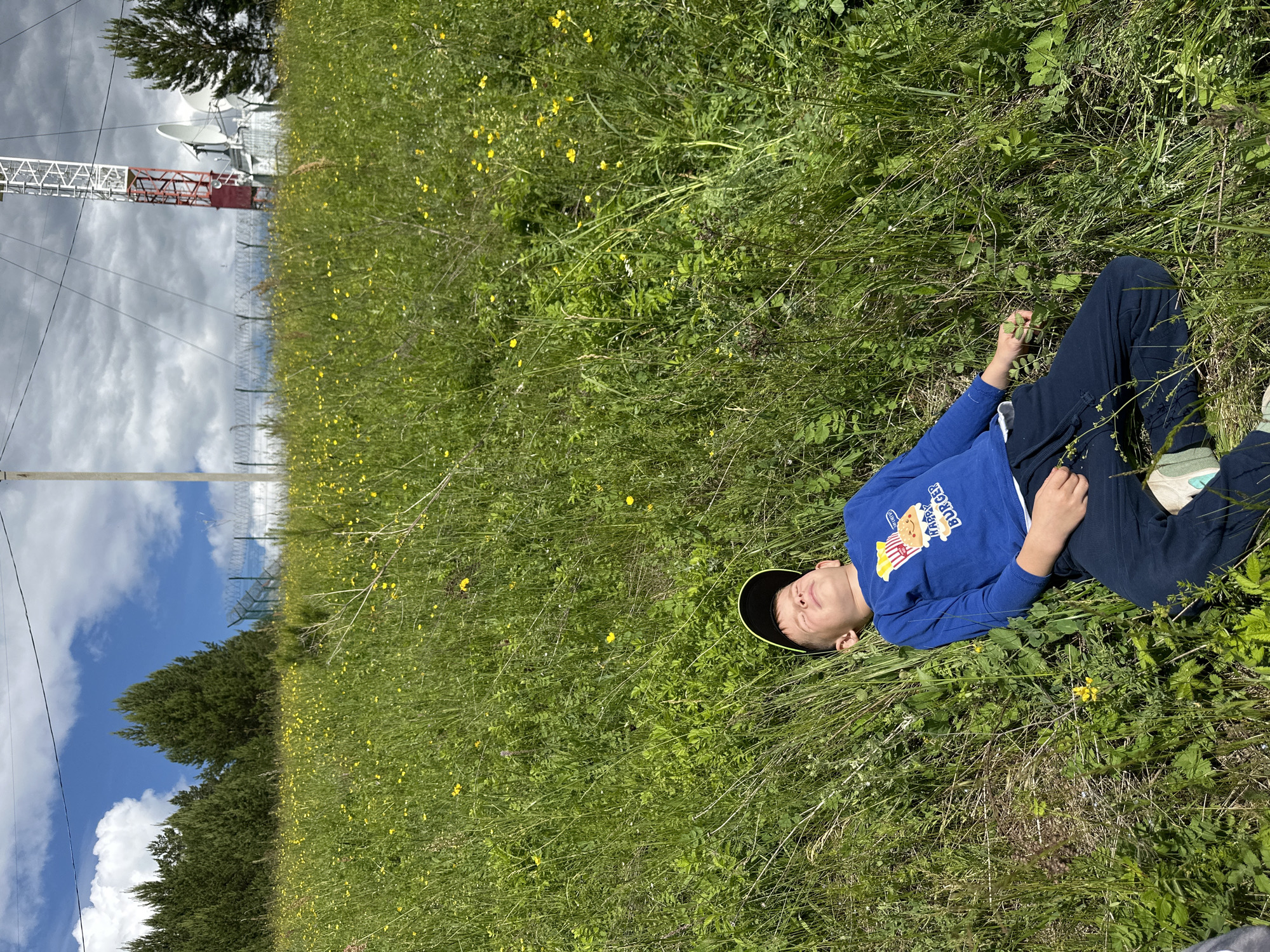 A young male child, estimated to be between 5 and 8 years old, lies supine in a vibrant green grassy field abundant with small yellow wildflowers under direct sunlight in the city of Альняш, Russia. The child's eyes are closed, and he appears to be resting or asleep. He is dressed in a blue long-sleeved shirt featuring a graphic of popcorn and a burger labeled "HAPPY BURGER," dark blue sweatpants with a white drawstring, and a black baseball cap with a green brim positioned backward on his head. His arms are relaxed at his sides, and light-colored sneakers with green accents are visible on his feet.
In the midground and background, the landscape rises. To the left, a dense cluster of coniferous trees is visible. Stretching across the horizon is a chain-link fence topped with barbed wire or razor wire, separating the natural field from an engineered site. Beyond the fence on the right, a tall red and white lattice communication tower is present, accompanied by multiple white satellite dishes and supporting structures. Overhead power lines and utility poles are also visible across the scene. The sky transitions from a clear blue with scattered white cumulus clouds on the left to a more overcast, grey cloud cover above the communication tower on the right.