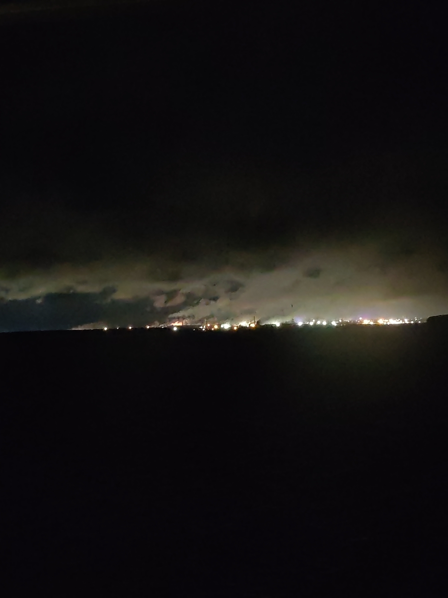 At night, a cityscape of Unknown, Unknown, is observed from a dark foreground. The skyline is illuminated by numerous scattered lights, suggesting a populated urban area. Above the city, a layer of clouds is present, partially obscuring the sky. The environment appears still and quiet, emphasizing the contrast between the dark foreground and the lit-up city. No people or specific objects are discernable in the immediate foreground.