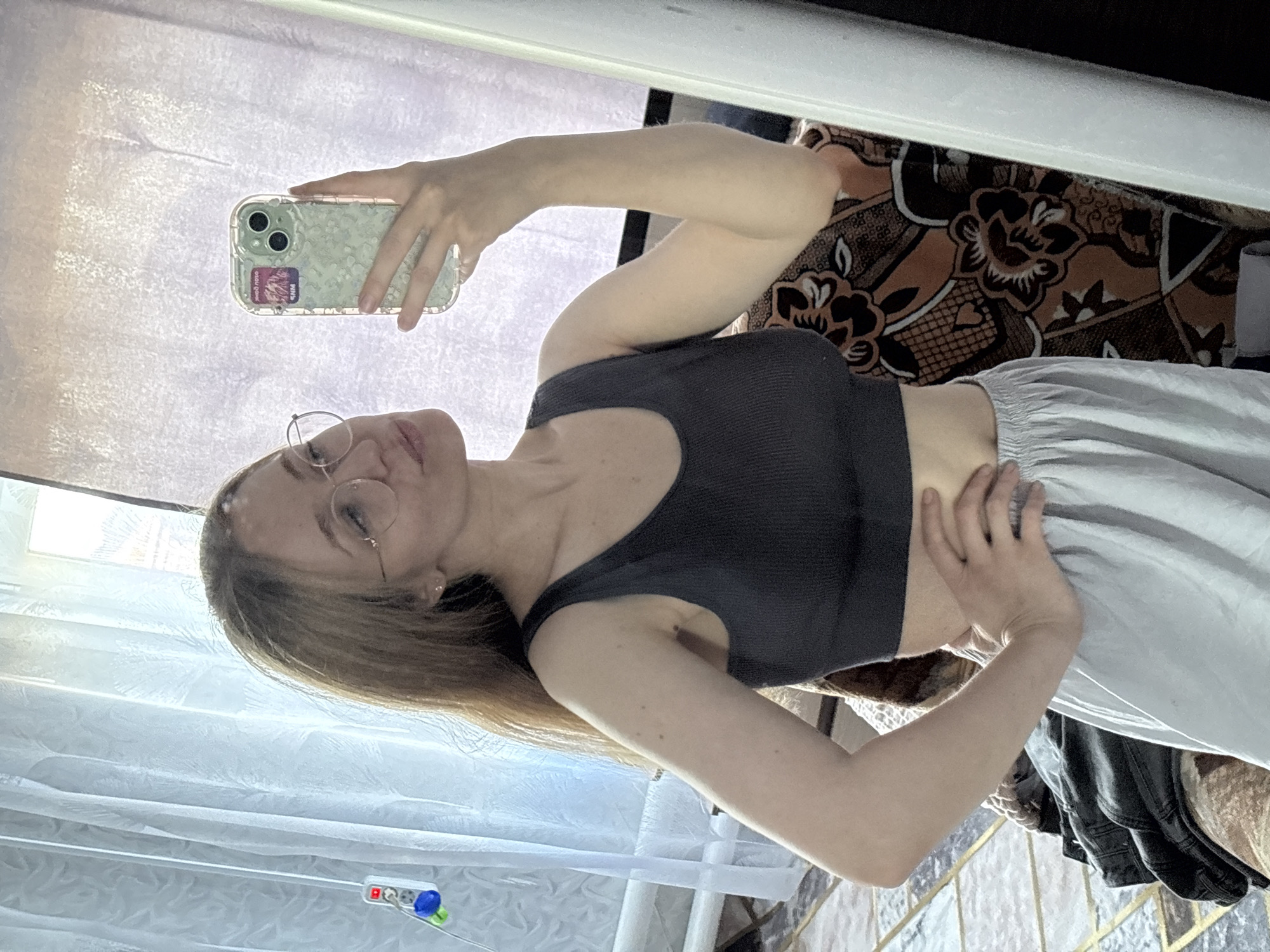 A young woman is captured in a mirror selfie, her body oriented vertically in the frame. She's wearing a dark, form-fitting tank top and light-colored sweatpants, with her right hand resting on her stomach. Her left arm is raised, holding a clear phone case with a floral pattern. She has blonde hair, glasses, and appears to be looking towards her phone, possibly at her reflection or the camera. The background is somewhat cluttered, with a patterned fabric visible behind her and a white curtain or blinds to her left. A power strip with multiple outlets and a blue object is visible near the bottom left of the image. The lighting suggests it might be daytime, with natural light coming from the left. The scene appears to be indoors, possibly a bedroom or living area.