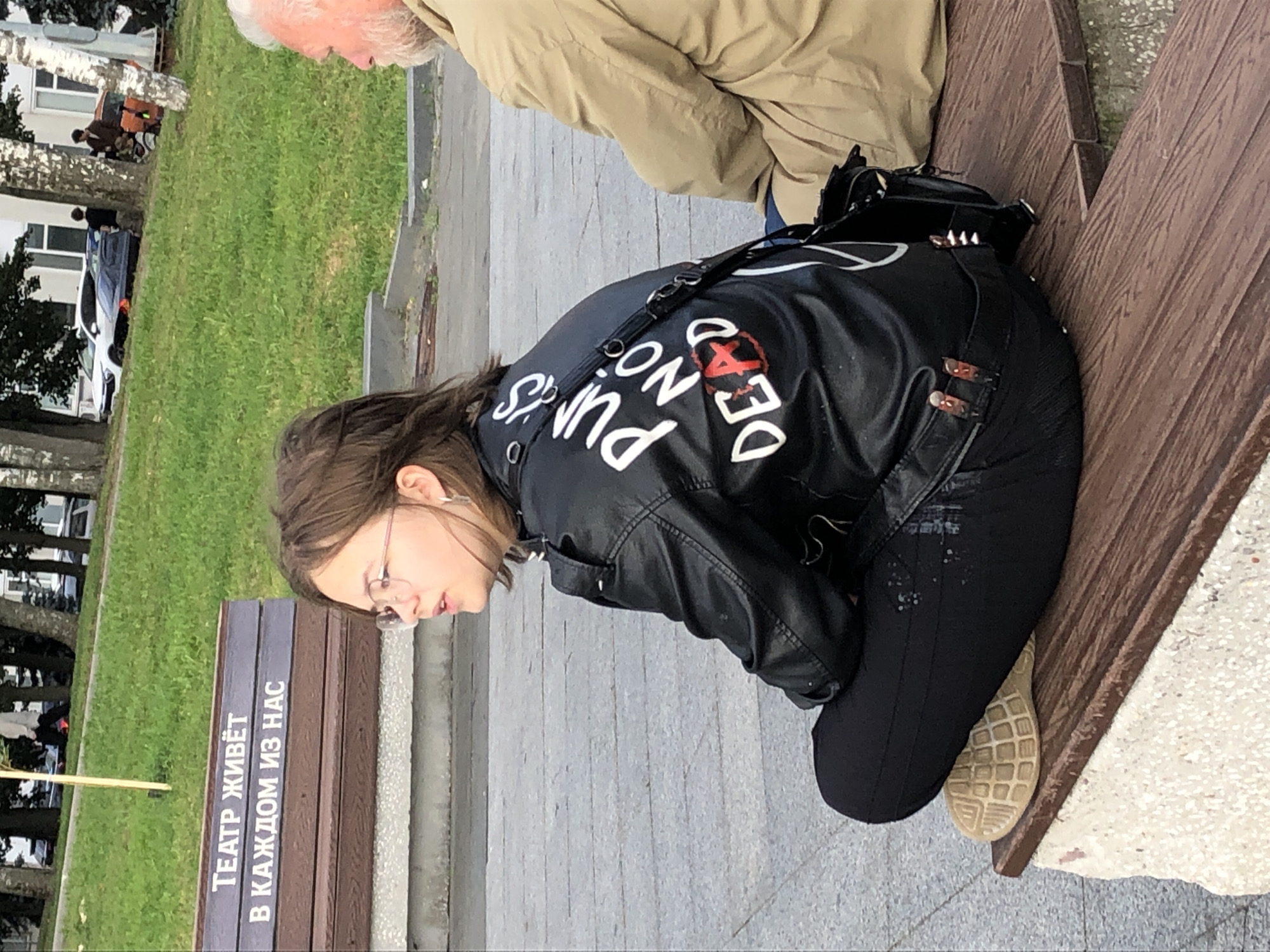 A young woman with short brown hair and glasses is sitting on a wooden bench. She is wearing a black leather jacket with white lettering that reads "Punks Not Dead" and black pants. To her right, an older man with white hair is also seated on the bench, his back is to the camera. The bench has text on it that reads "ТЕАТР ЖИВЁТ В КАЖДОМ ИЗ НАС" which translates to "The theater lives in each of us". The background shows a grassy slope with trees and buildings, suggesting an outdoor urban setting. The woman is looking to the right, away from the camera.