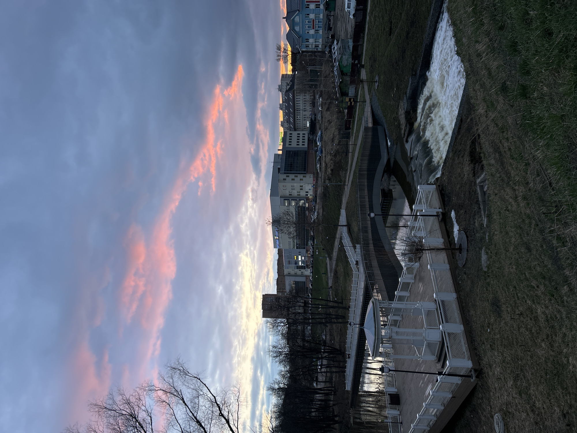 The image shows a cityscape at sunset in Klin, Russia. A cascade of water flows from a raised concrete structure into a waterway. A viewing platform with white metal railings and lampposts overlooks the water. Beyond the platform, a row of bare trees lines the water's edge. Several buildings of varying heights and architectural styles are visible in the background. Cars are parked near some of the buildings. The sky is filled with gray clouds, with streaks of pink and yellow indicating the sunset. Grass covers the embankment, transitioning from green to brown due to the season.