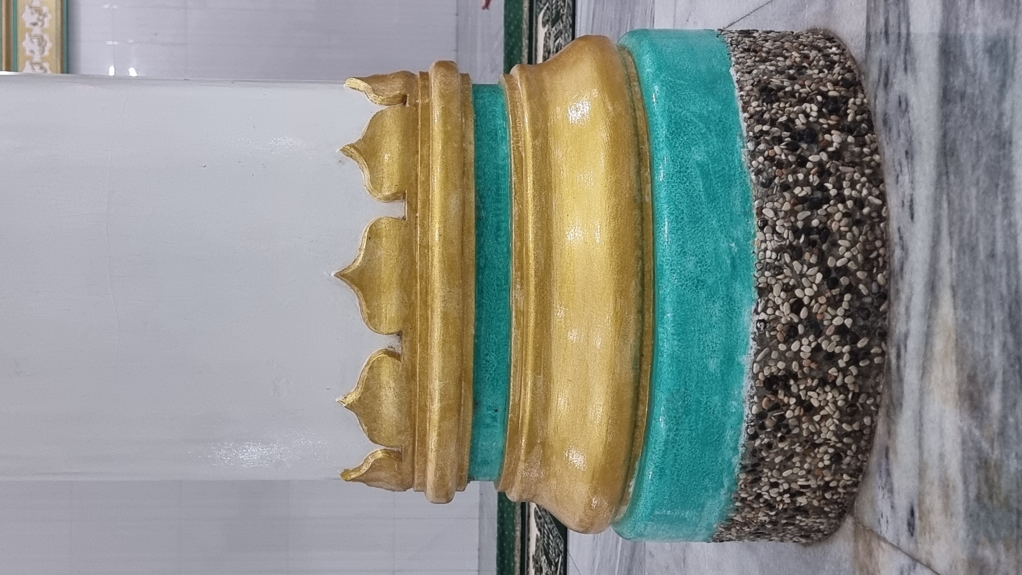 The image shows the base of a white pillar with gold and turquoise decorative elements. The bottom section of the pillar is covered in small, colorful pebbles. The pillar is situated on a marble floor, with a patterned carpet visible in the background, suggesting an indoor environment. The context provided about Lapang Timu, Indonesia, indicates this is likely a decorative pillar within a public space or building. No people are clearly visible in the frame.